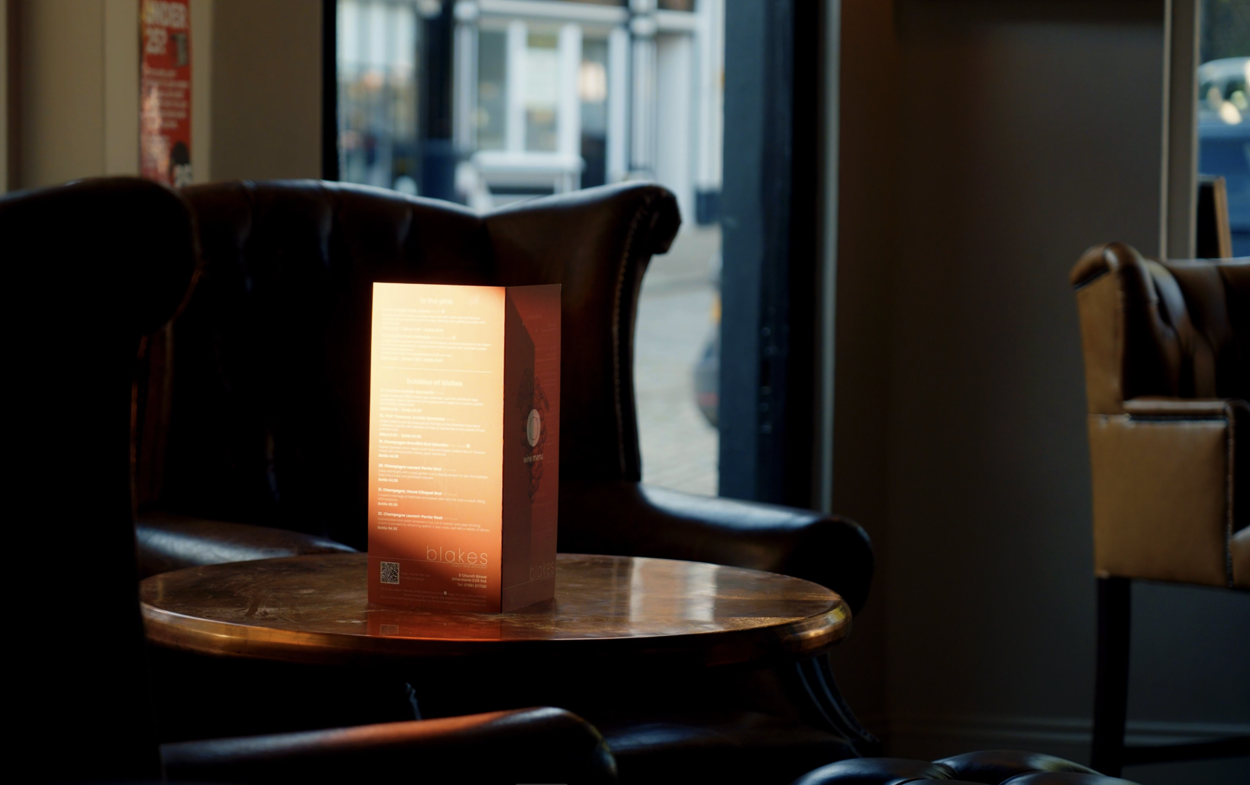 A lit menu card on a round wooden table inside a dimly lit cafe, with leather chairs and a partially open door showing an outdoor view of a street and parked cars.