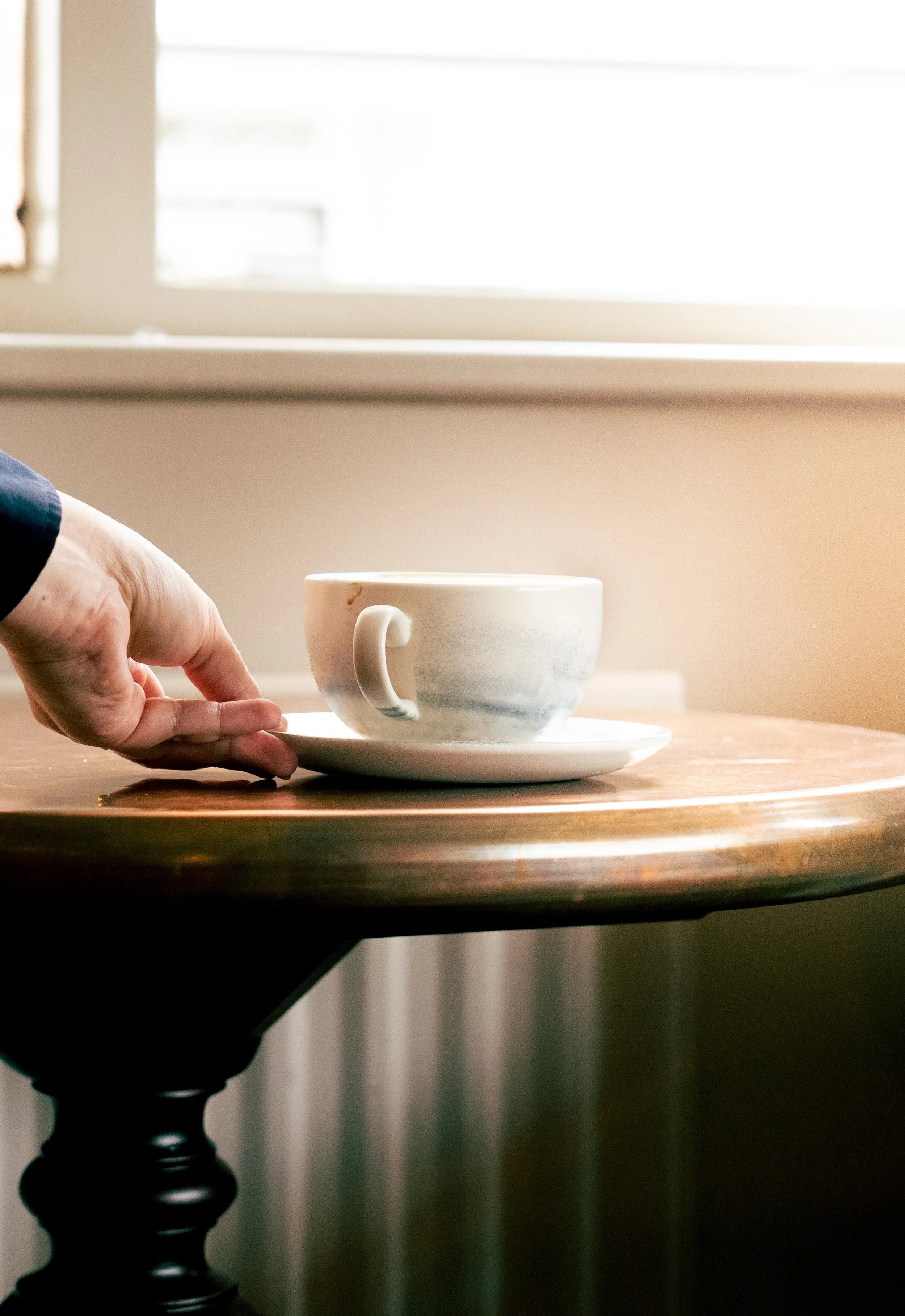 A hand reaches for a ceramic cup on a saucer on a wooden table near a window with sunlight.