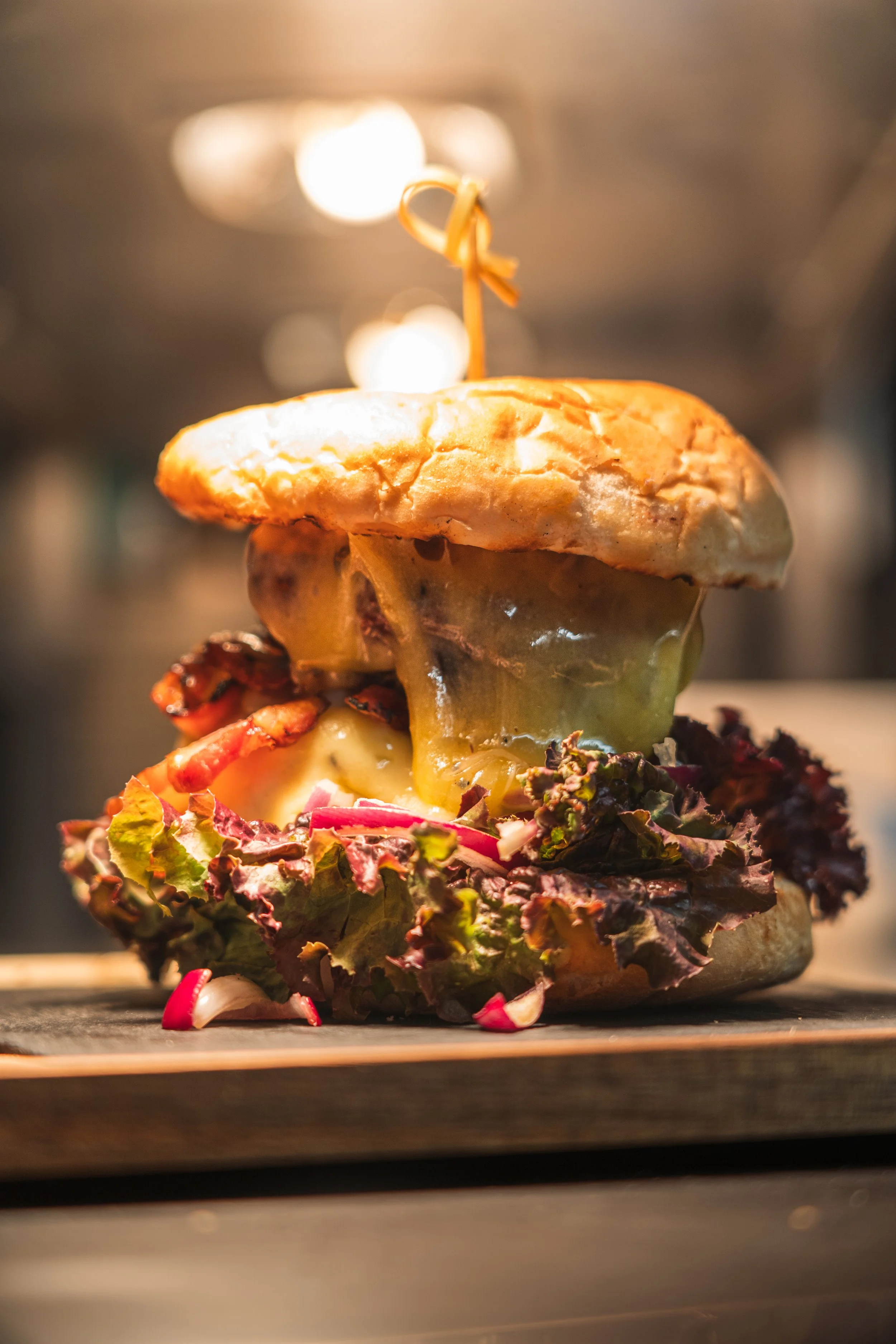 Close-up of a cheeseburger with lettuce, bacon, cheese, and a bun, skewered with a toothpick, on a wooden surface.