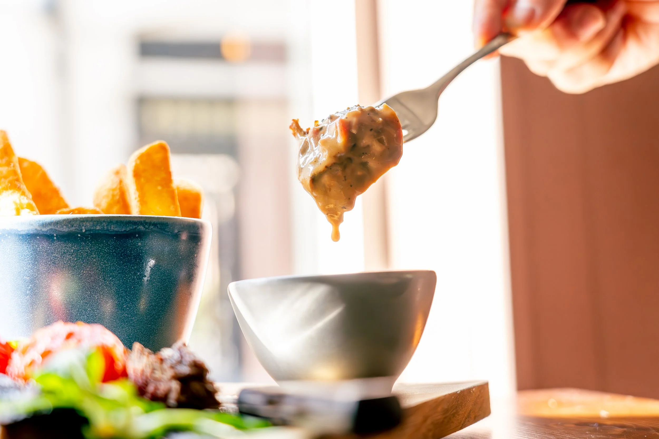 Person scooping thick, creamy gravy onto a small white bowl at a breakfast table with fried potatoes and fresh vegetables in the foreground.