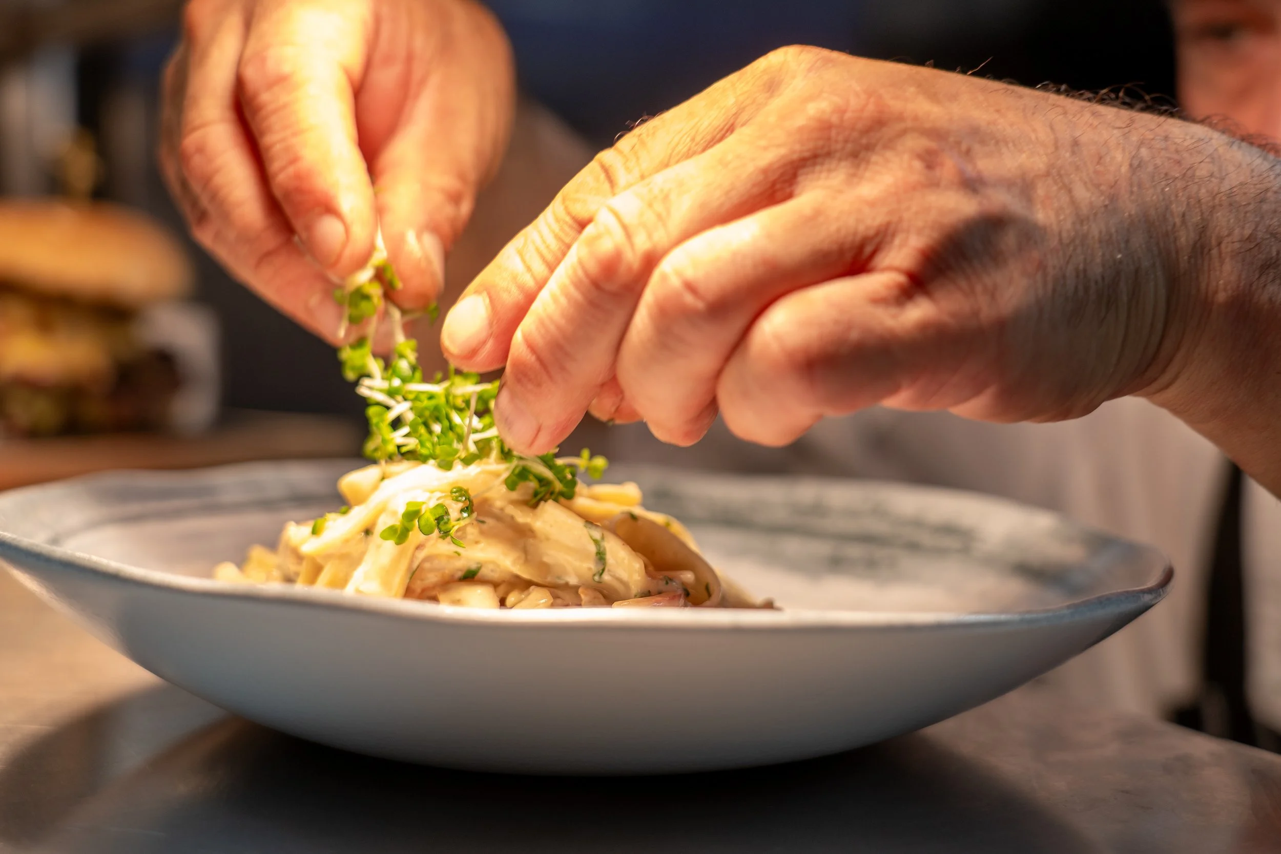 Hands garnishing a plated pasta dish with microgreens in a restaurant setting.