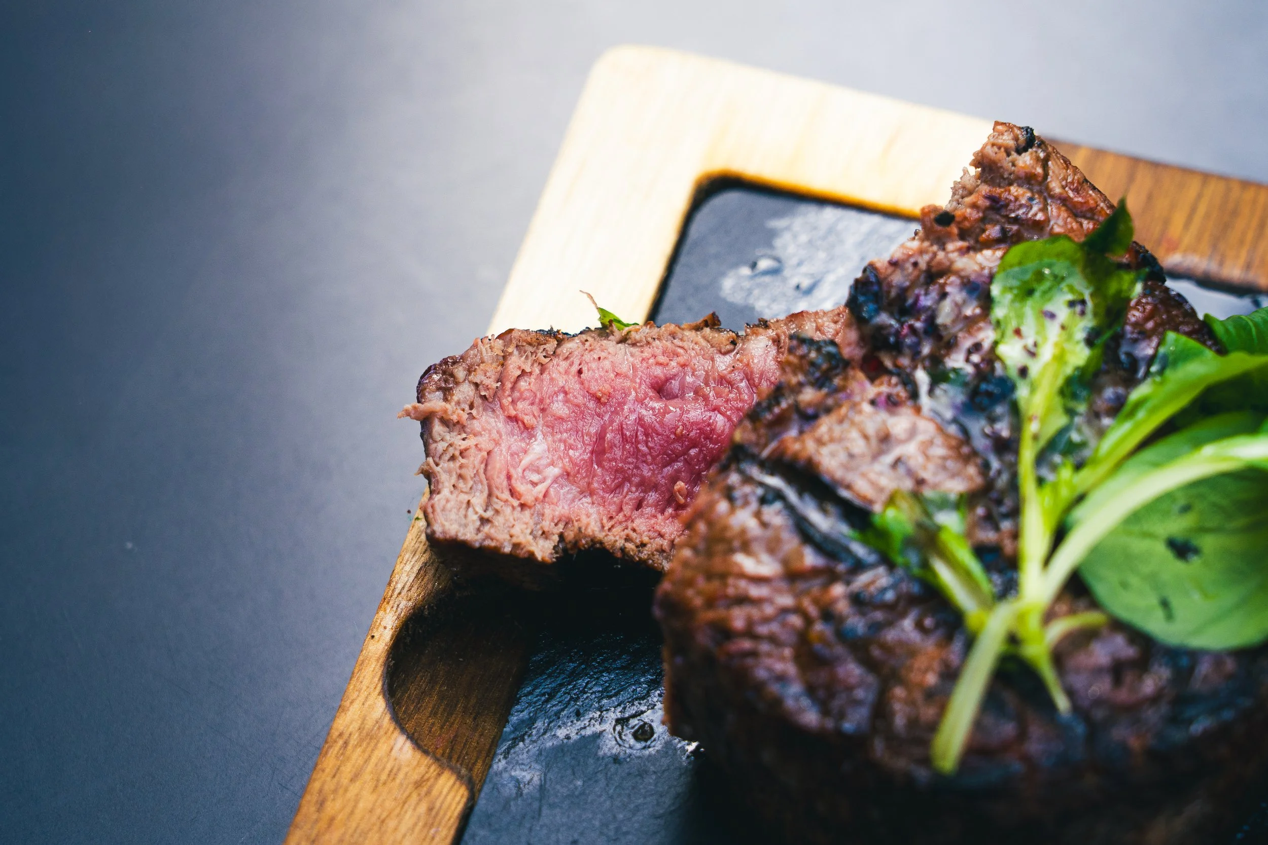 Close-up of a sliced beef wellington on a wooden serving board, with rare pink interior and green garnish, on a dark surface.
