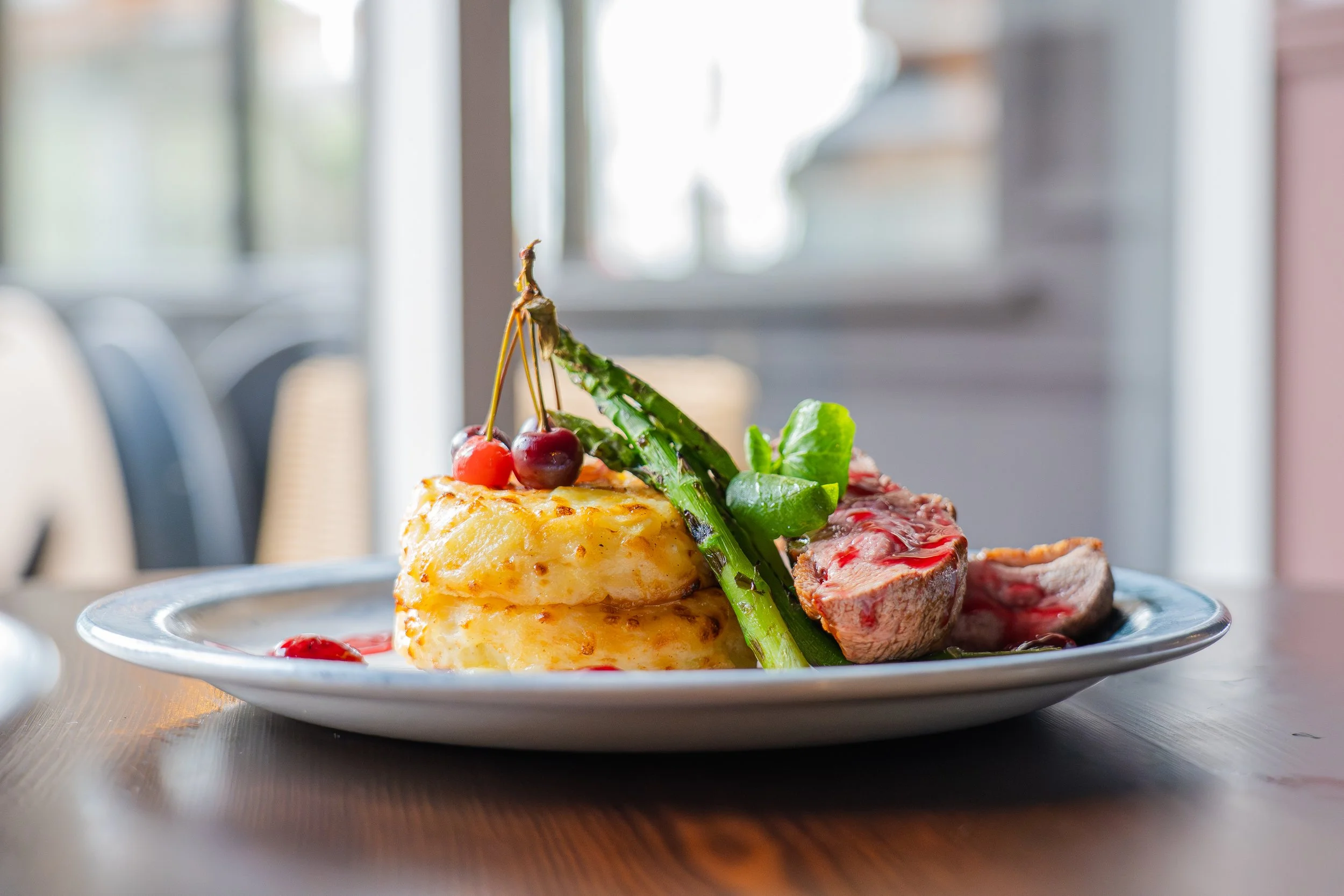 A plate of food featuring a stack of mashed potatoes topped with cherries, accompanied by grilled asparagus, a piece of steak with sauce, and some greens on a wooden table in a bright, modern setting.