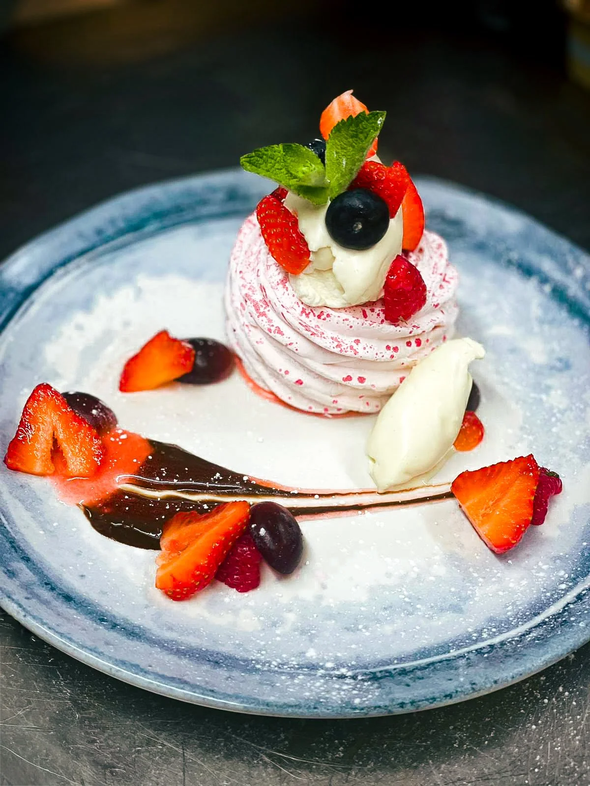 A dessert with sliced strawberries, blueberries, a mint leaf, and a swirl of pink cream, garnished with a white chocolate piece, on a blue plate dusted with powdered sugar.