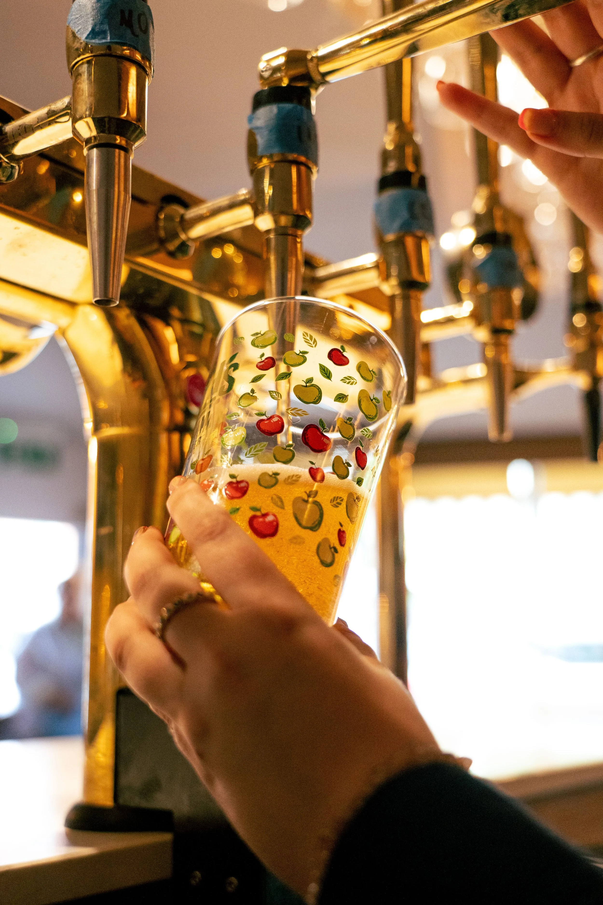 A person is pouring beer from a tap into a glass with decorative red and green fruit patterns. The bar tap is gold-colored, and the background is blurred with warm lighting.