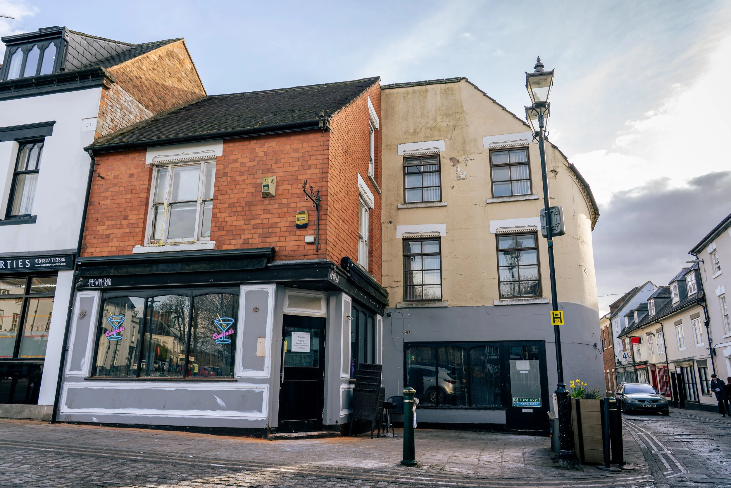 Street view of three connected buildings, with a bar on the ground floor, including a white facade, large corner window, and signs for cocktails, aluminum windows, a street lamp, and parked cars.