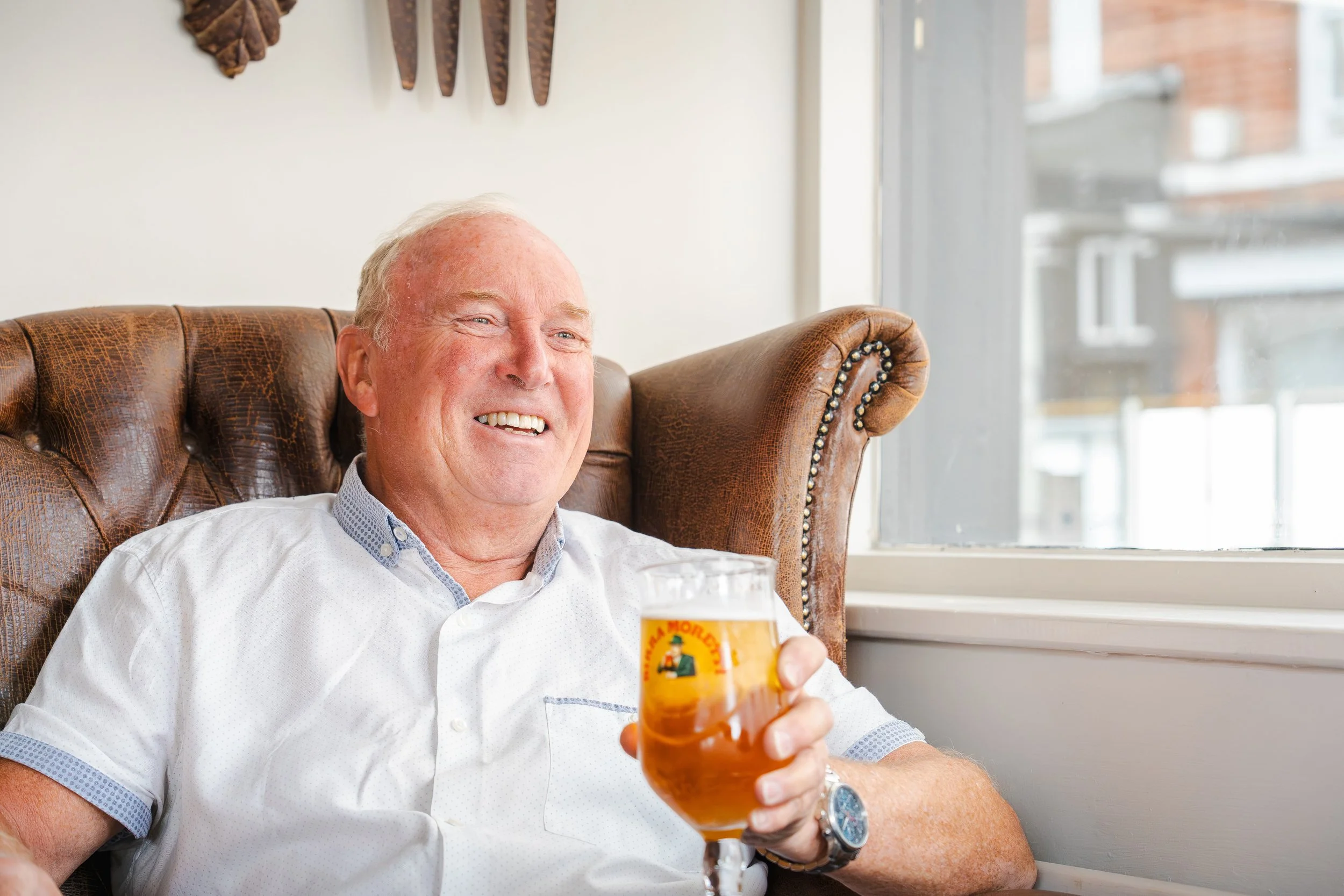 An elderly man with white hair and a white button-up shirt sitting in a leather armchair, holding a glass of beer, smiling while looking out the window.
