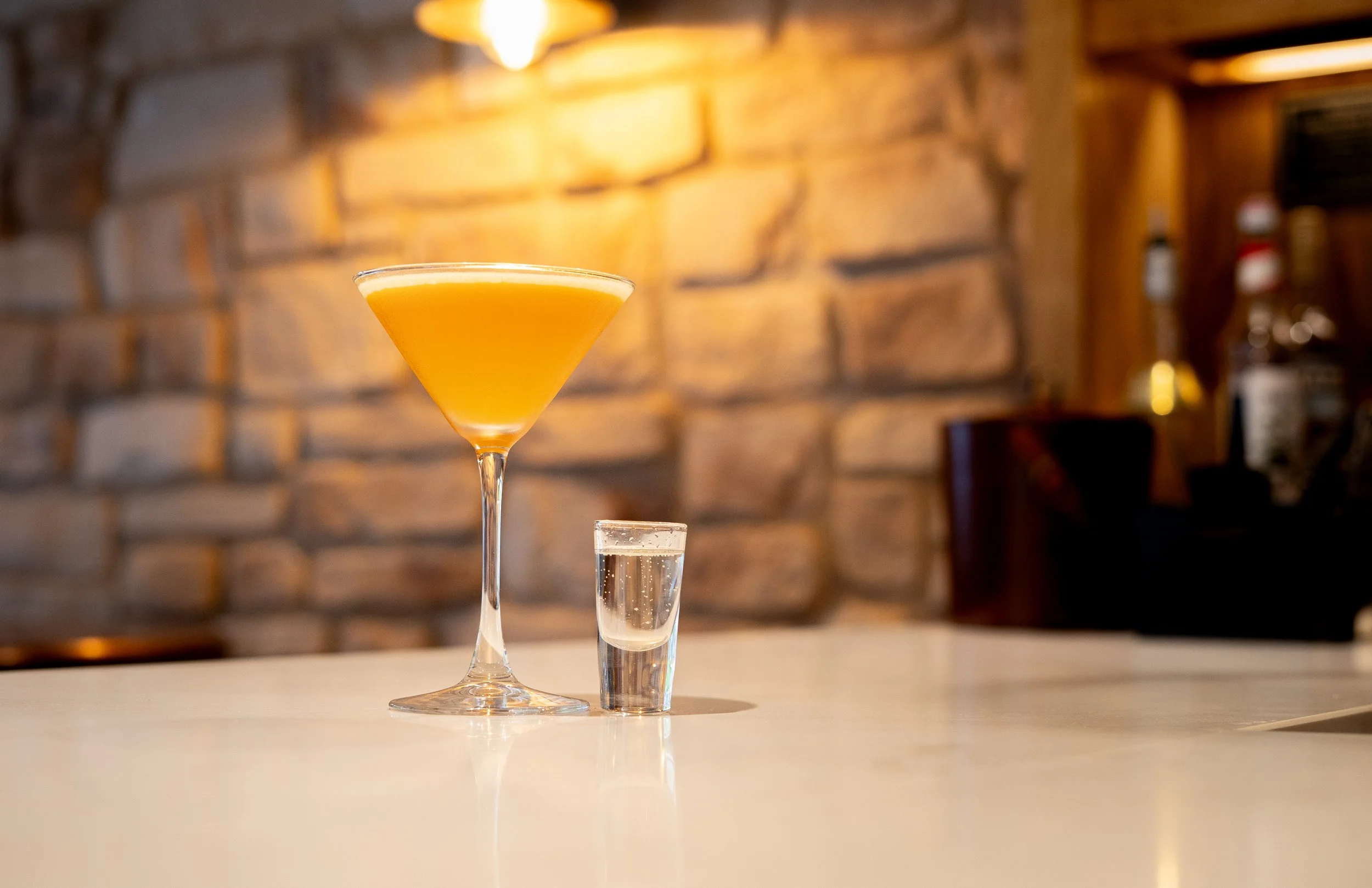 A cocktail glass with orange drink and a shot glass with clear liquor on a white bar counter, against a background of a brick wall and wooden shelves with bottles.