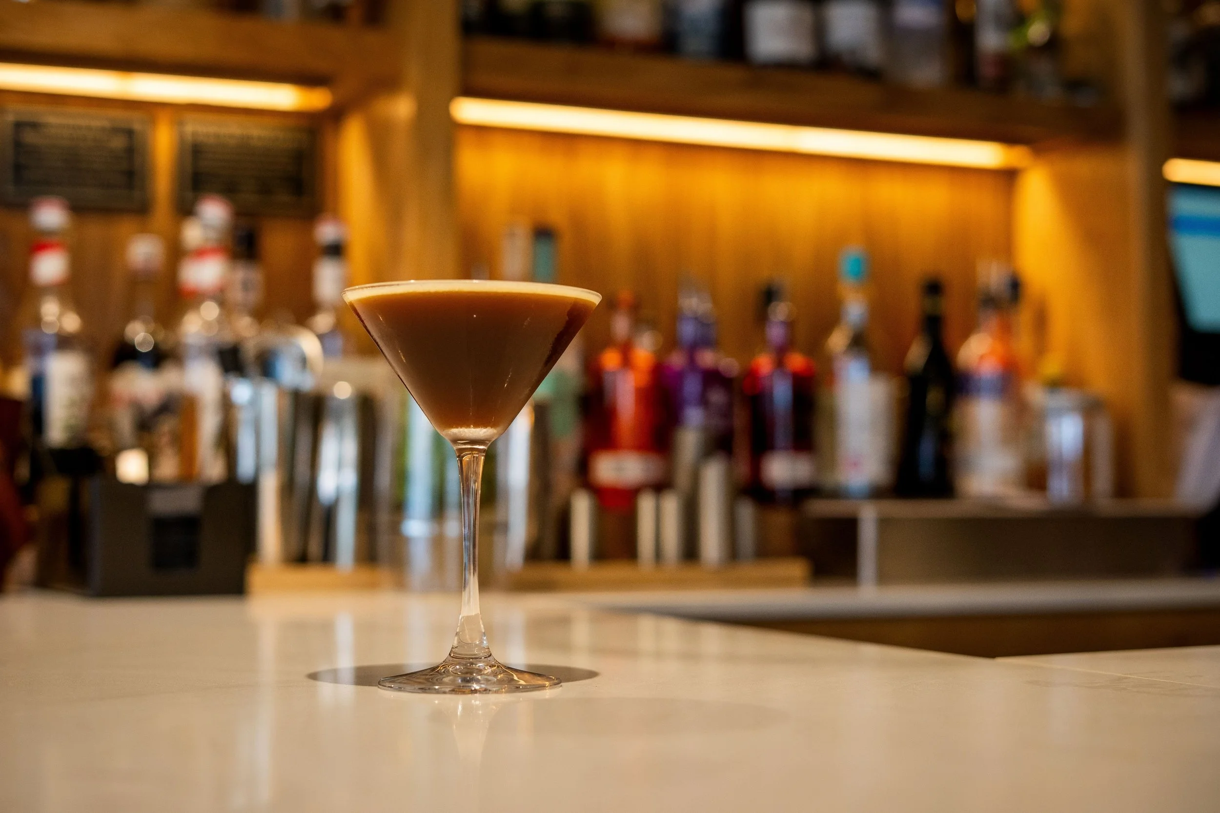 A cocktail glass filled with a dark brown drink on a bar counter, with bottles of alcohol and bar tools in the background.