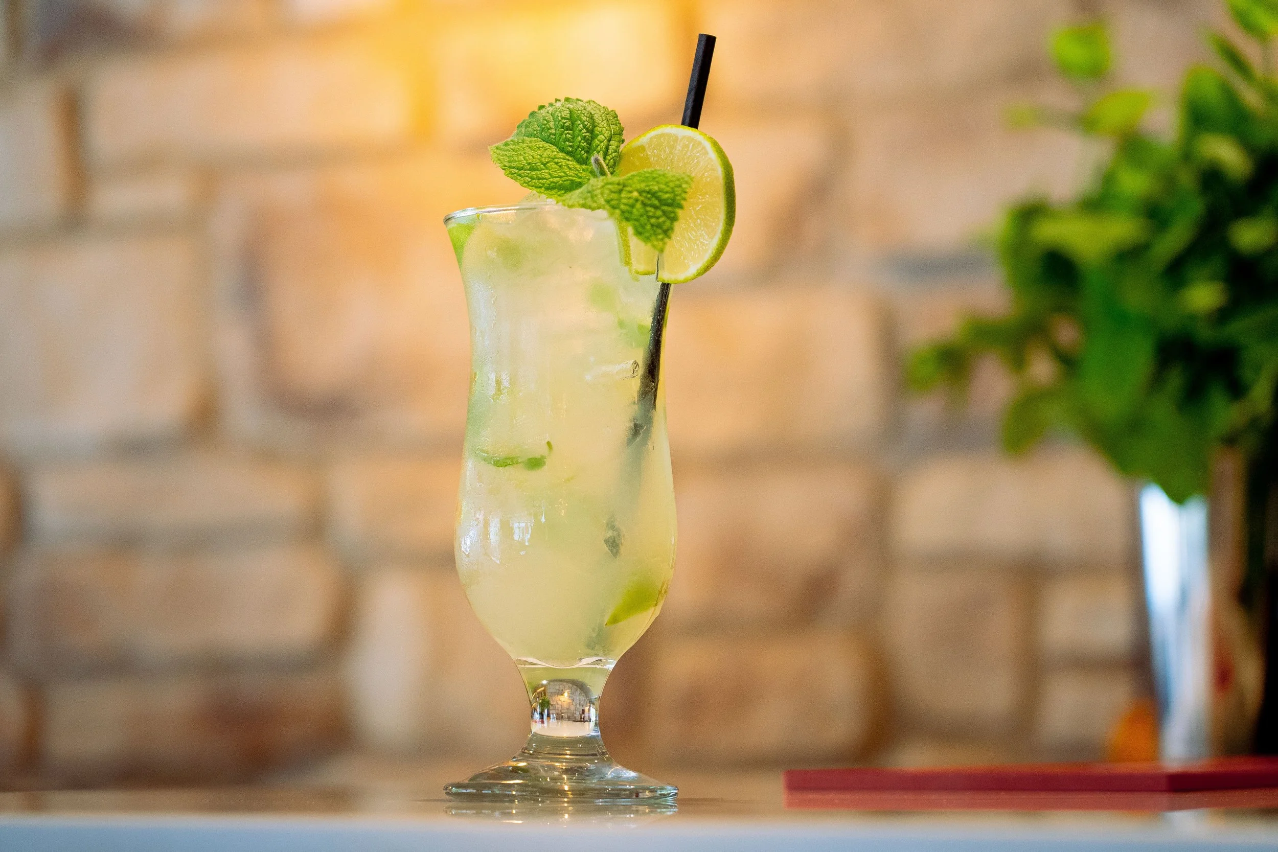 A tall glass of mojito cocktail with lime slices, mint leaves, ice, and a black straw on a table, with a brick wall in the background.