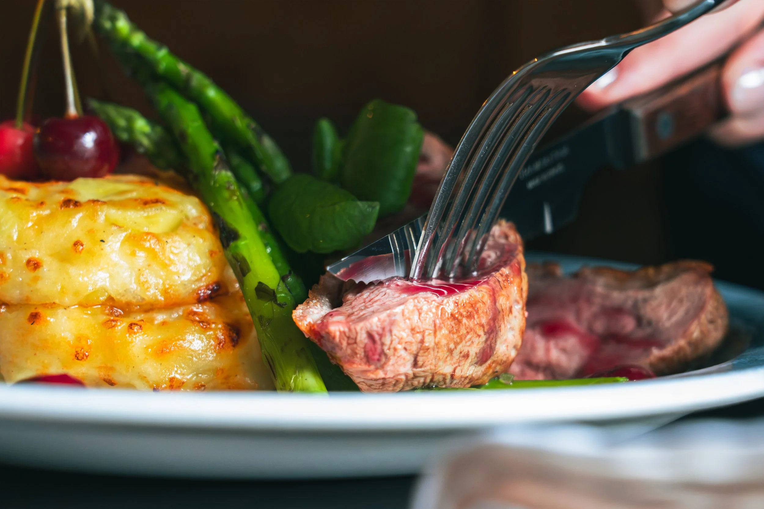 Close-up of a plate with a baked cheesy dish, grilled green asparagus, cherries, and a piece of cooked steak with a fork piercing it.