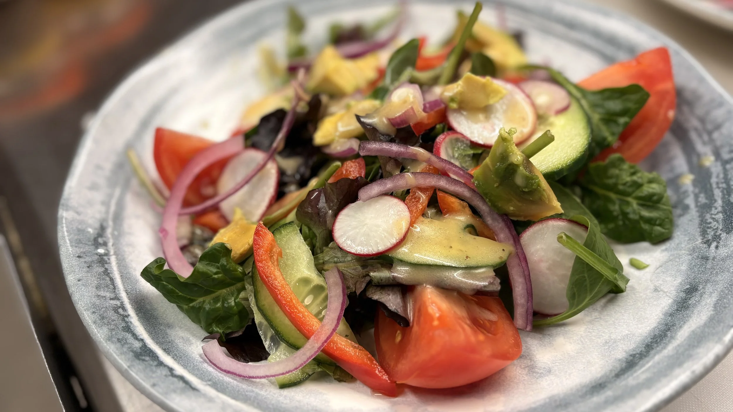 Fresh mixed vegetable salad with sliced radishes, cucumbers, tomatoes, red bell peppers, avocado, spinach, and red onions served in a white bowl.