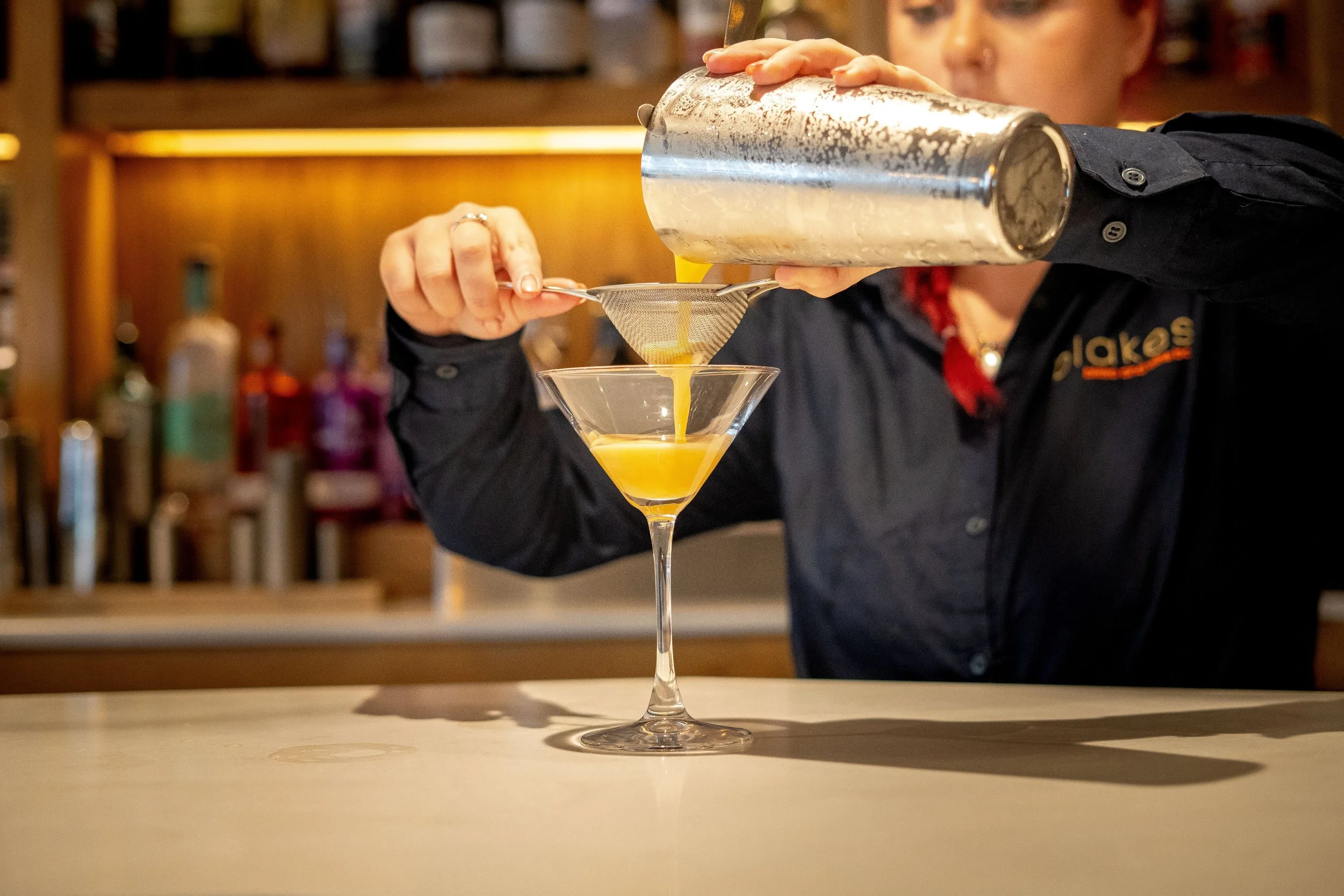 A bartender or mixologist pouring a yellow cocktail through a strainer into a martini glass at a bar.