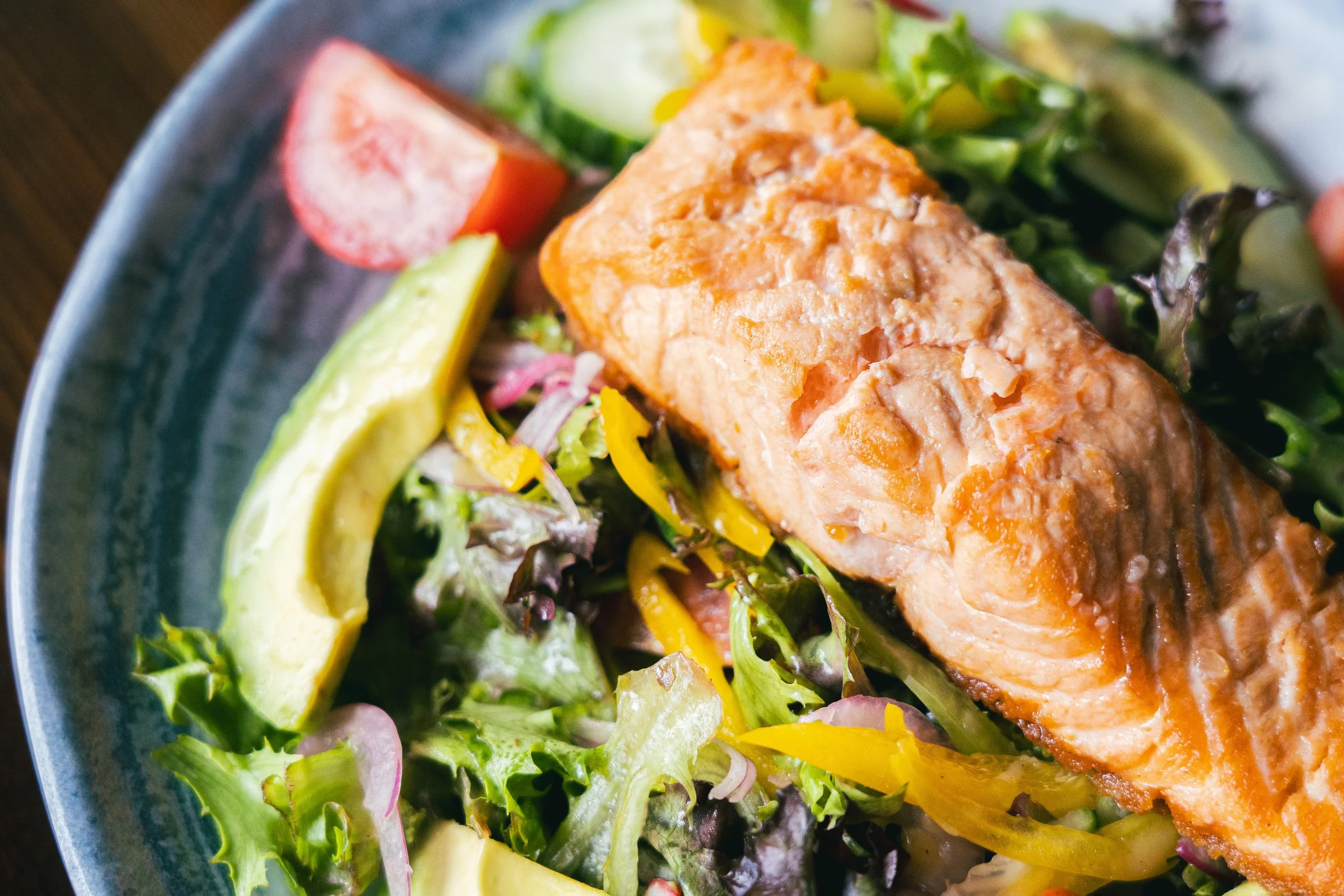 Salmon fillet on a bed of mixed greens, sliced avocado, cherry tomato, and yellow bell pepper