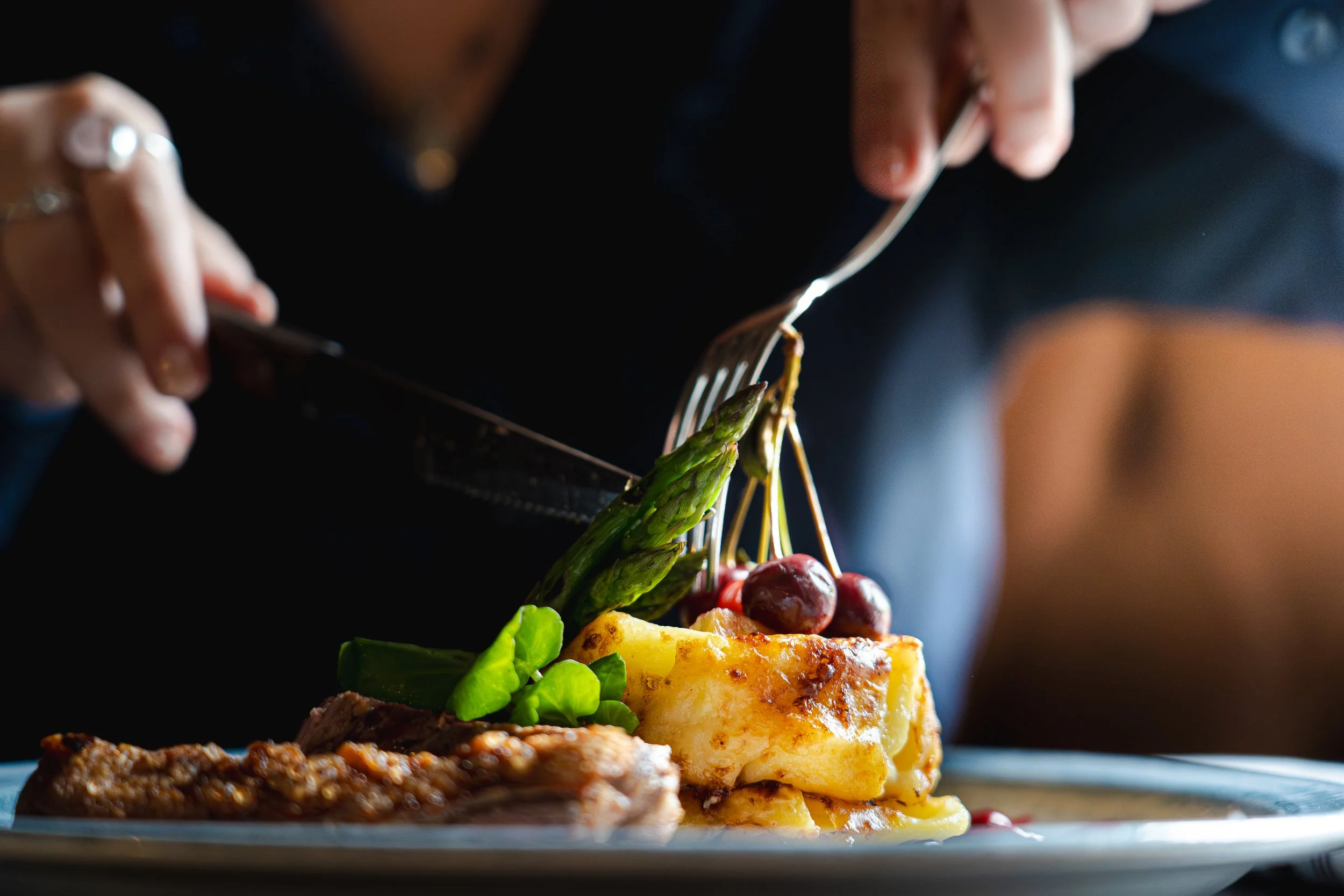A person is cutting into a plate of food with a fork and knife, revealing lasagna, asparagus, cherries, and greens.