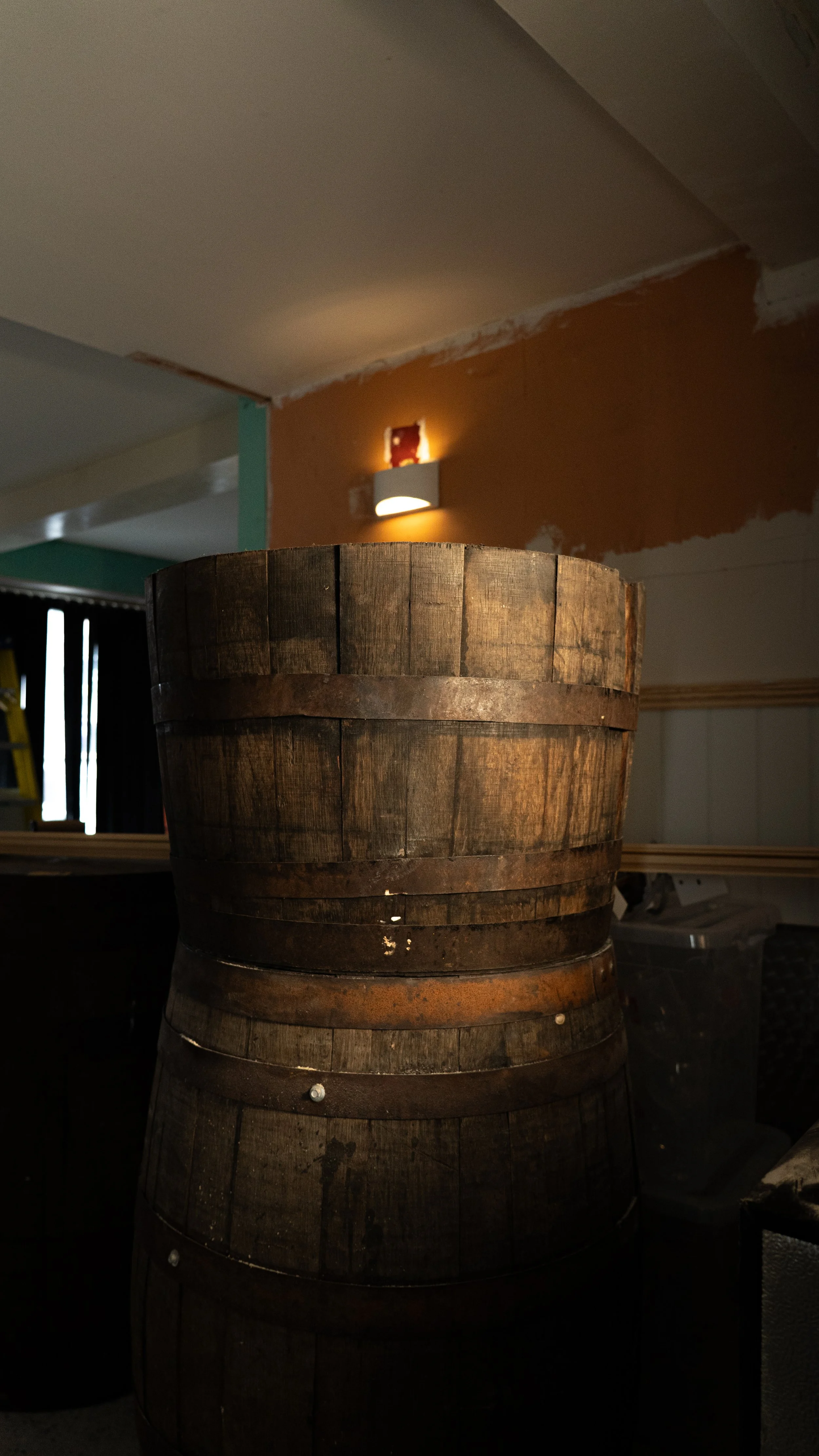 A large wooden barrel with metal bands in a dimly lit room, with a wall light and a partially painted wall in the background.