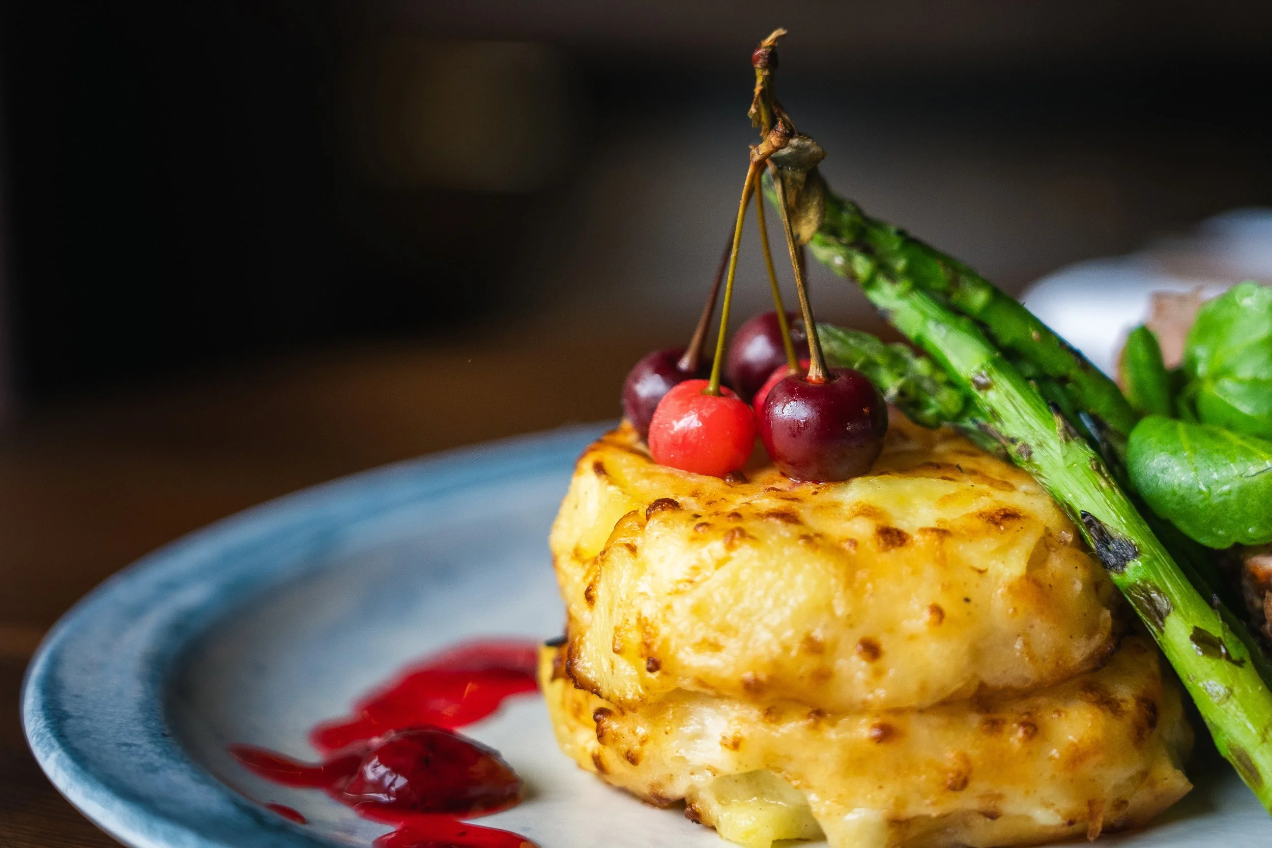 A close-up of a grilled chicken breast topped with cherries, grilled asparagus, and green leafy vegetables on a blue-rimmed plate, with a red sauce drizzle.