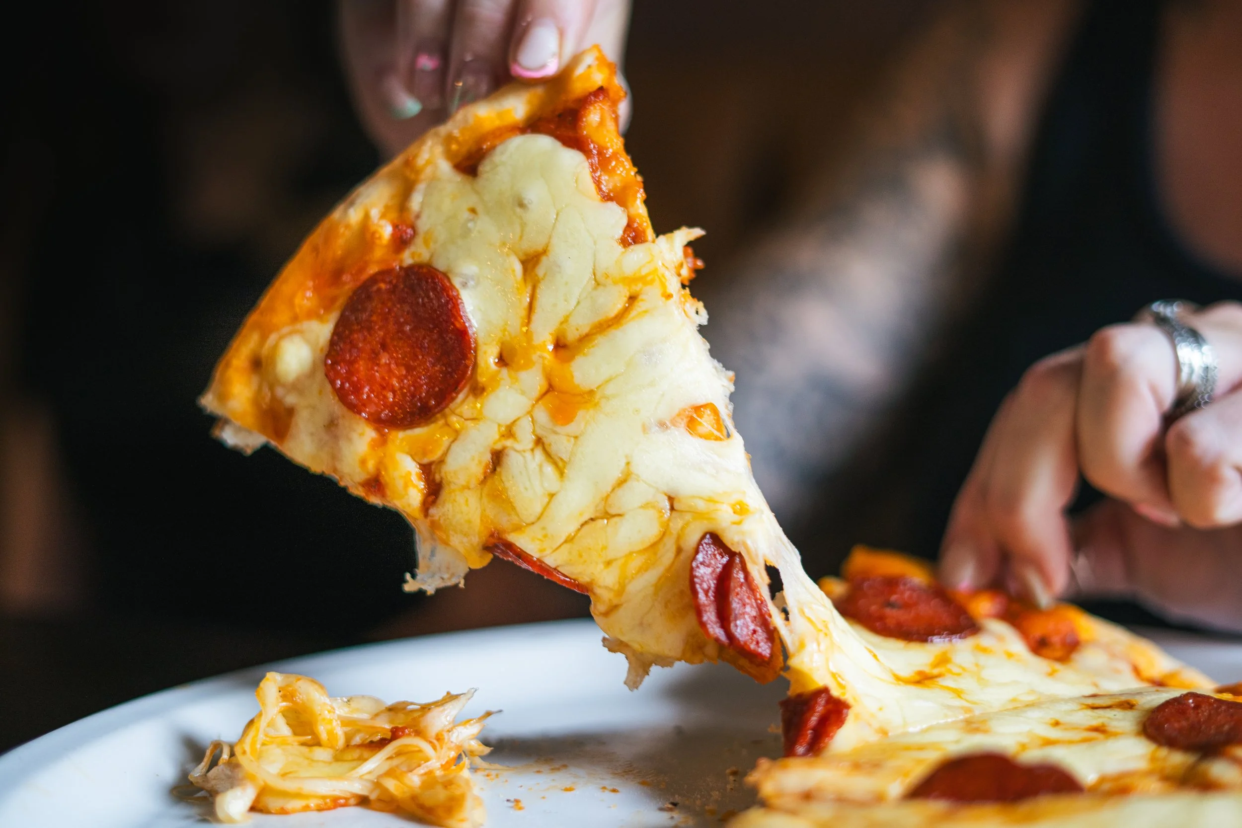 Person lifting a slice of pepperoni pizza from a white plate, with melted cheese and pepperoni toppings visible.