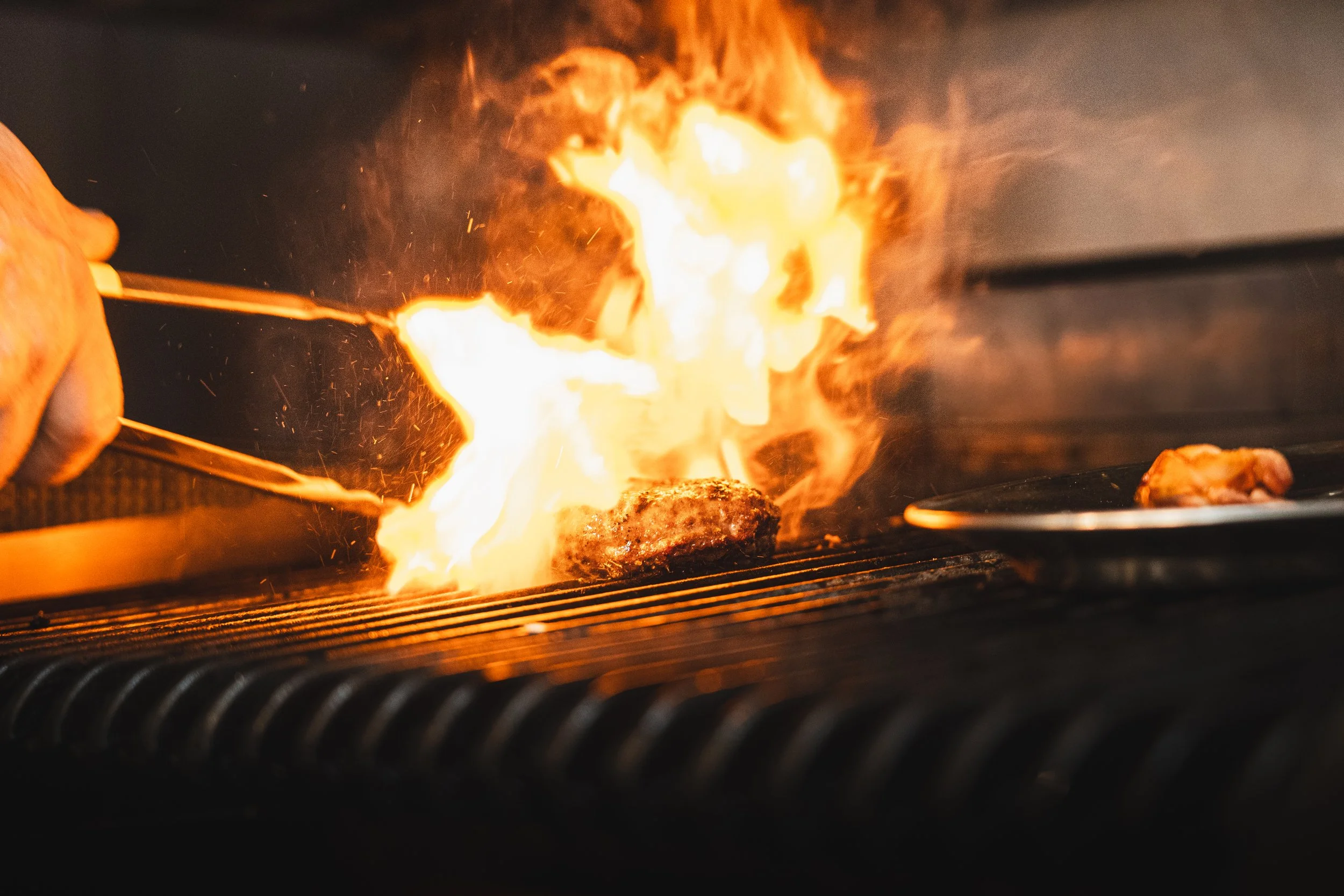 Chef flambéing a steak on a grill with flames and fire in a kitchen.