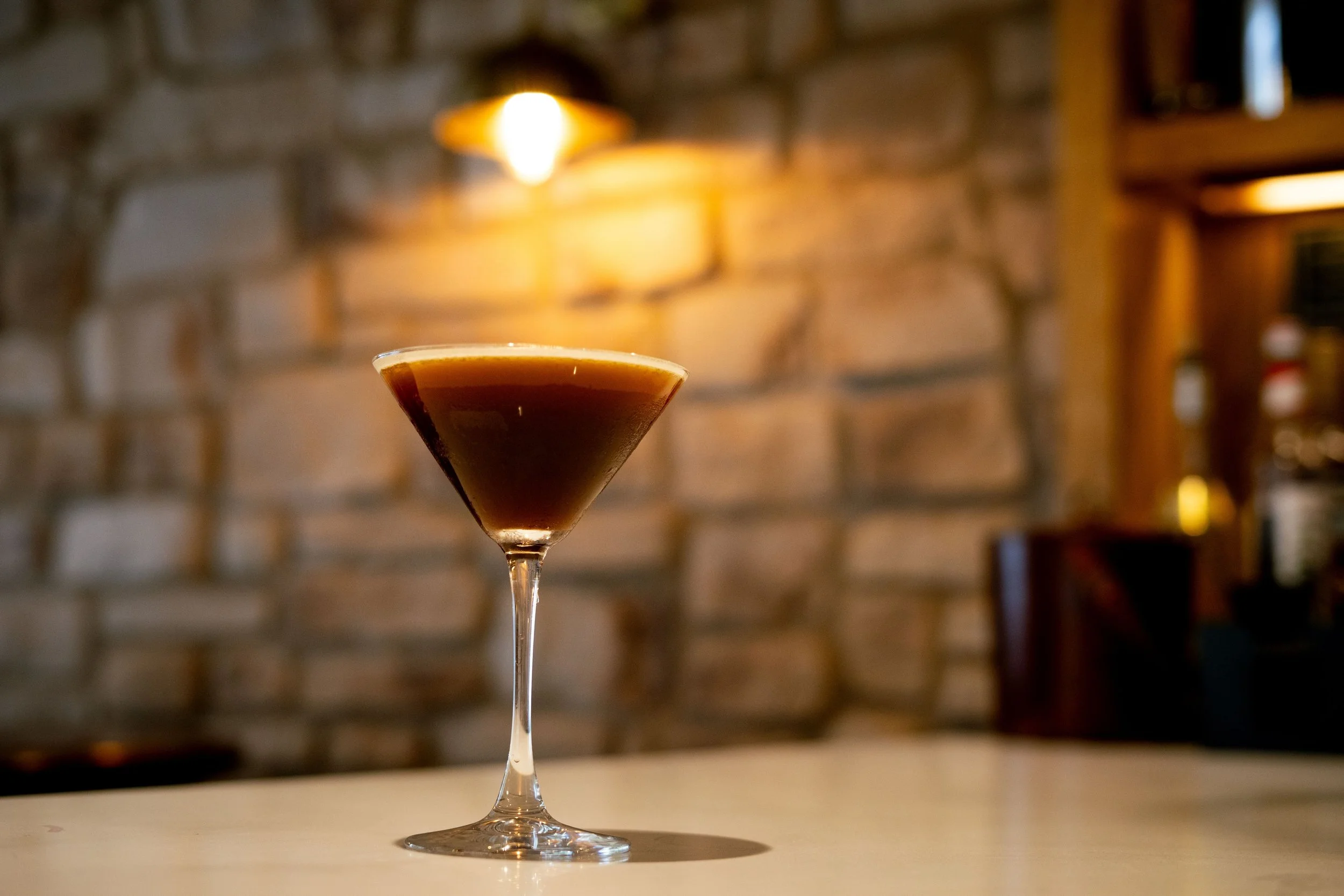 A cocktail in a martini glass on a bar counter, with a blurred brick wall background and warm lighting.
