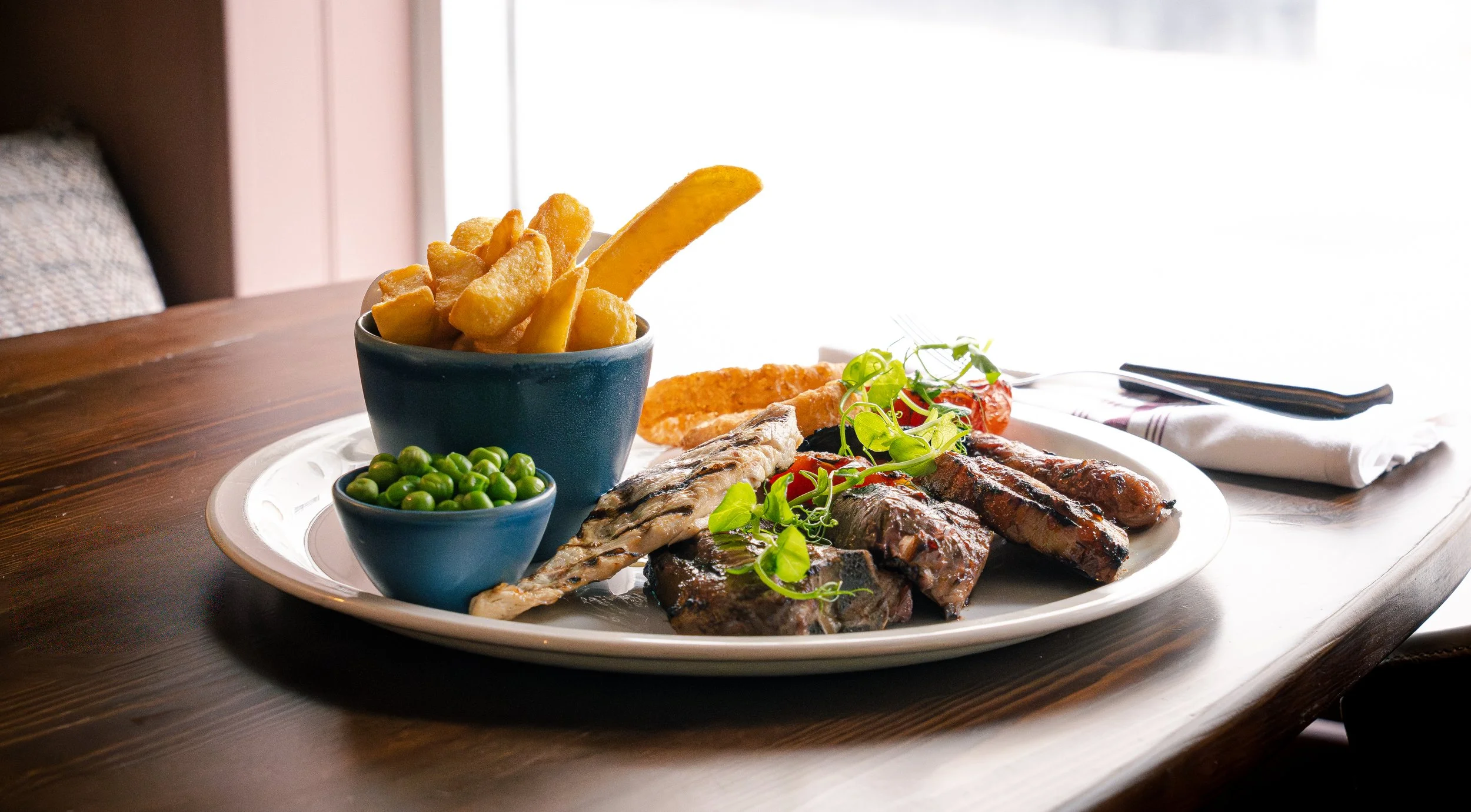 A large white plate of grilled meats, vegetables, and sides on a wooden table near a window, including grilled ribs with greens, fish, cherry tomatoes, and a basket of fries with a side of green peas.