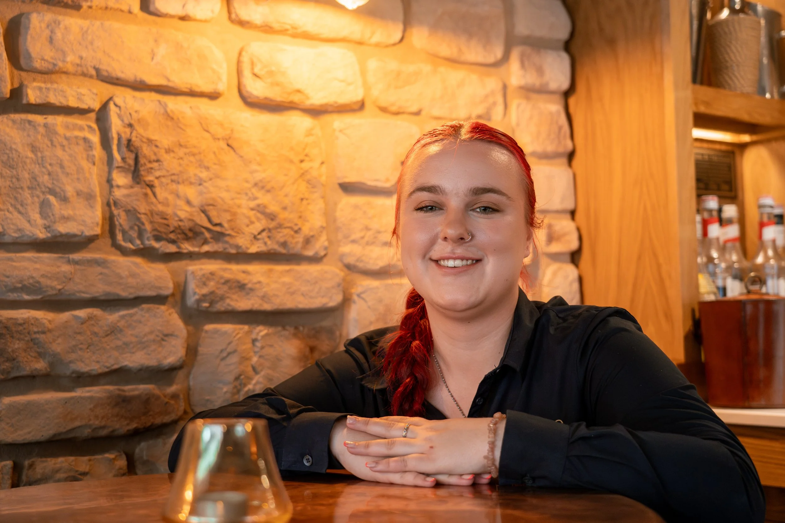 A young woman with red hair smiling, sitting at a table in a cozy restaurant with a stone wall background.