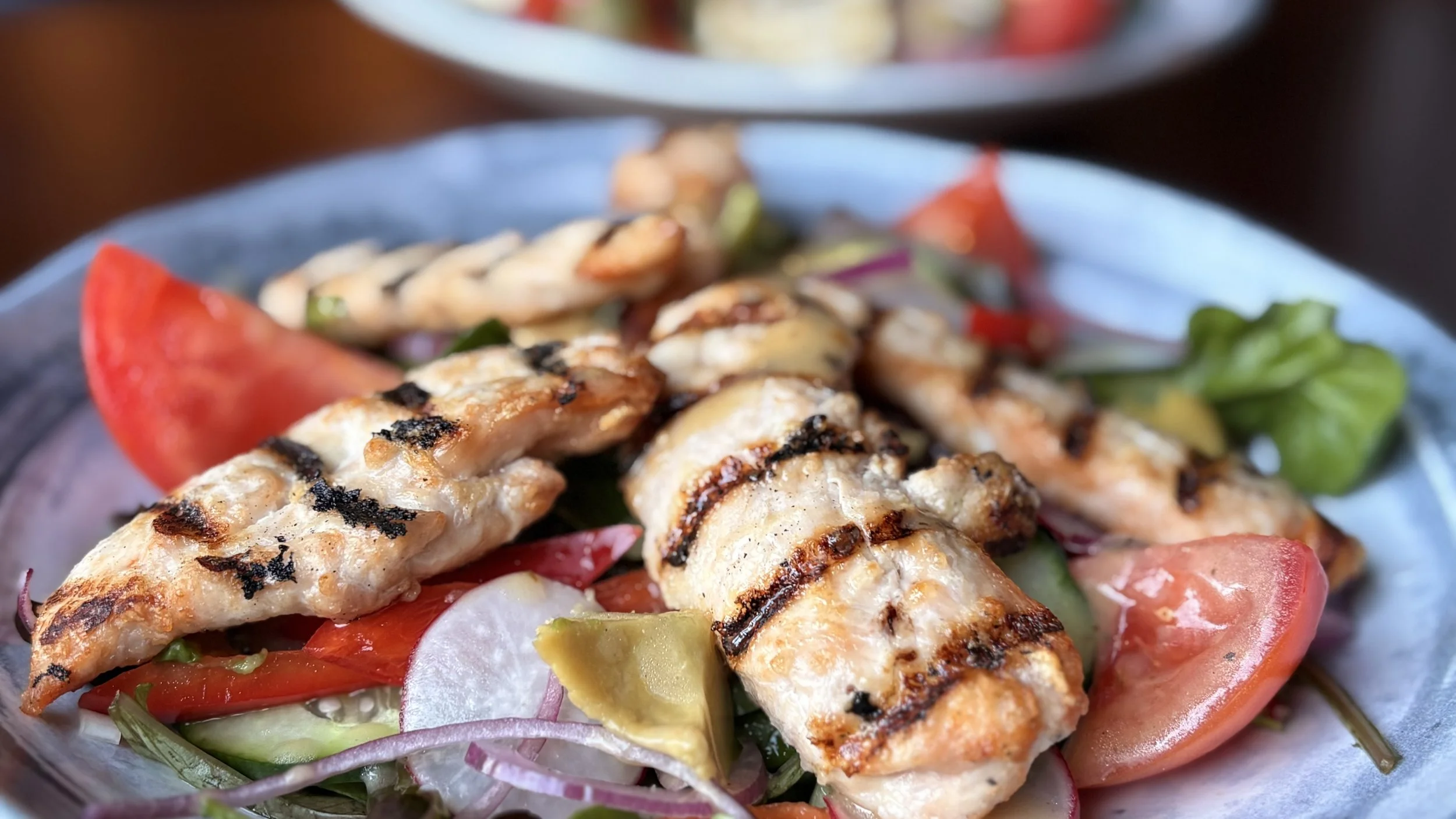 Grilled chicken salad with tomatoes, cucumbers, radishes, and greens on a blue plate.