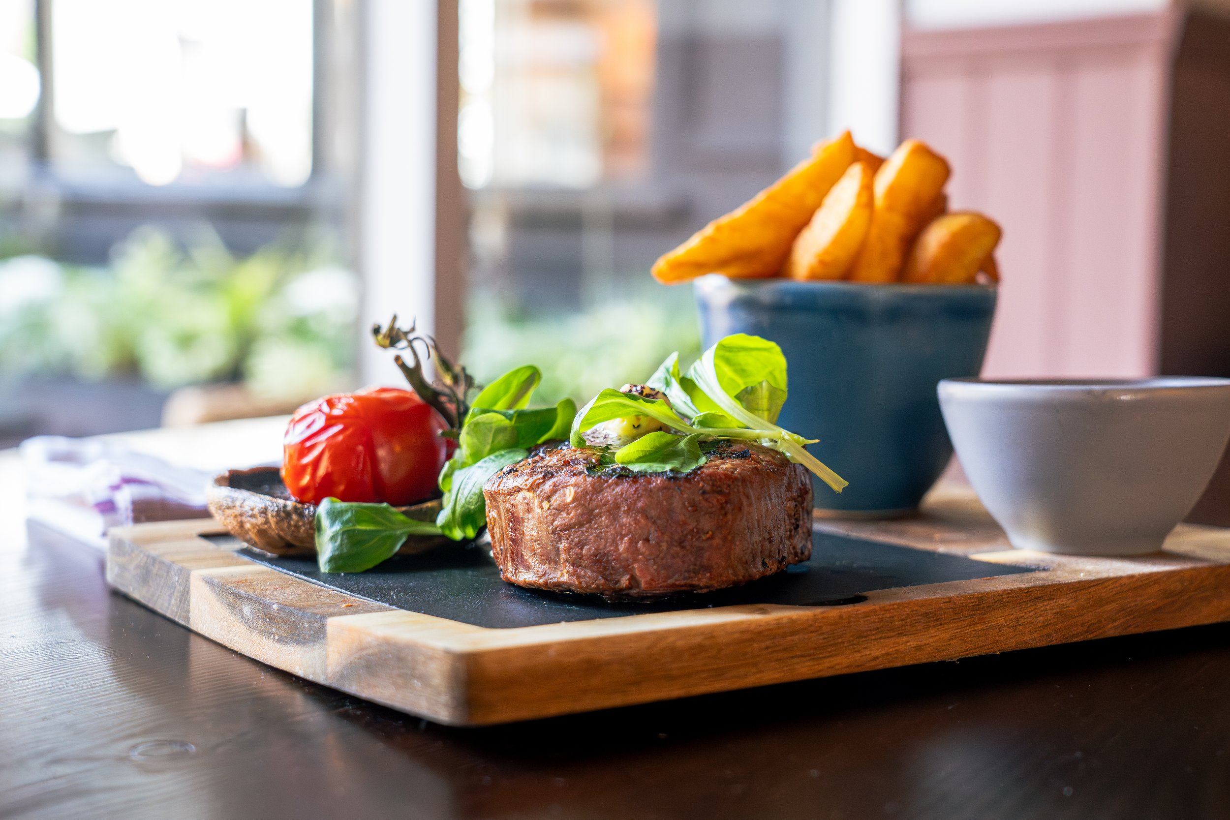 A cooked steak topped with fresh greens, served with a cherry tomato and grilled mushroom on a black slate plate, alongside a bowl of potato fries and two empty white bowls on a wooden serving board.