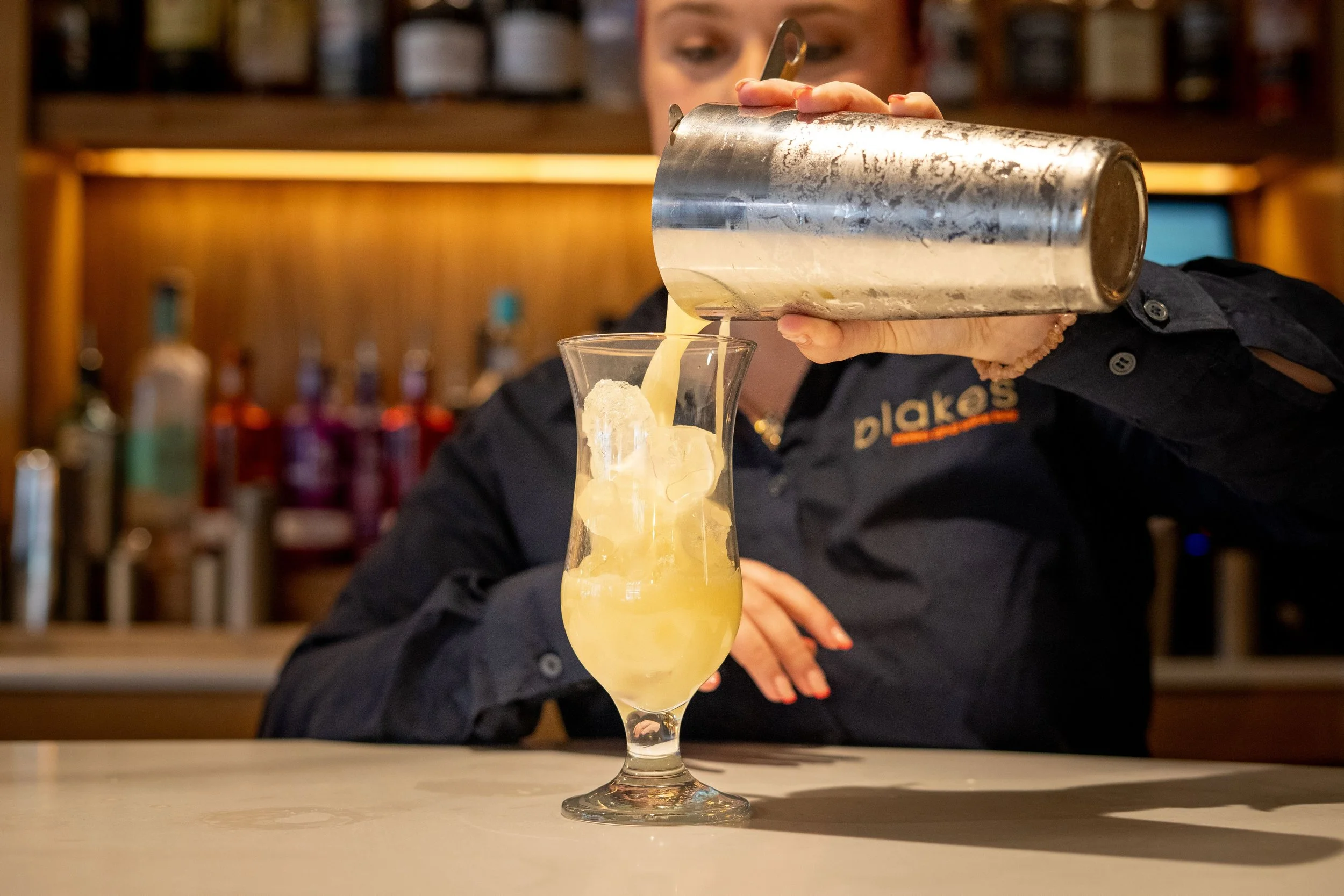 Bartender pouring a yellow cocktail into a glass with ice at a bar.