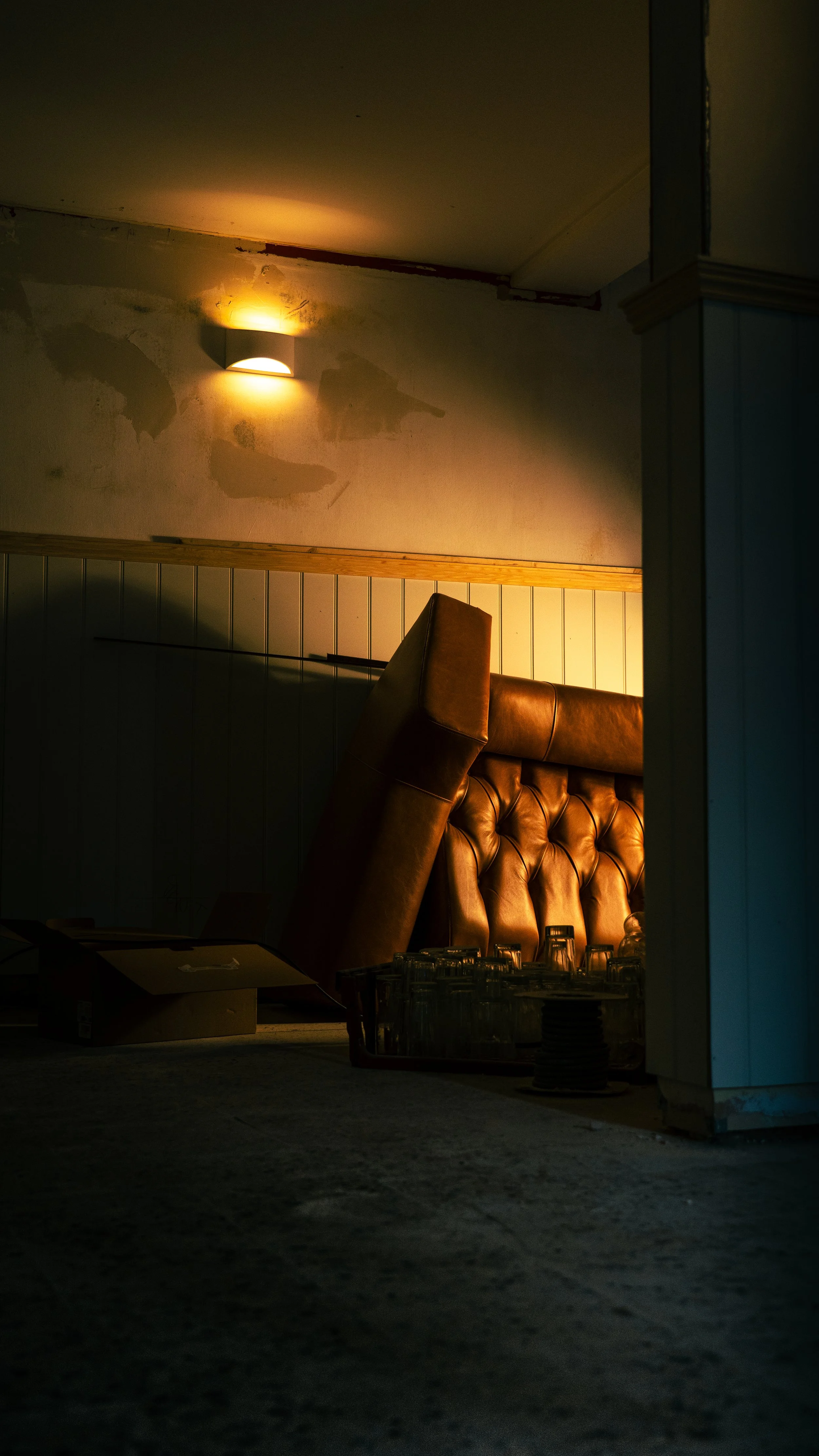 A dimly lit room with a single wall lamp on the wall, casting shadows, and a large brown leather padded headboard leaning against the wall, surrounded by glassware and an open cardboard box on the floor.