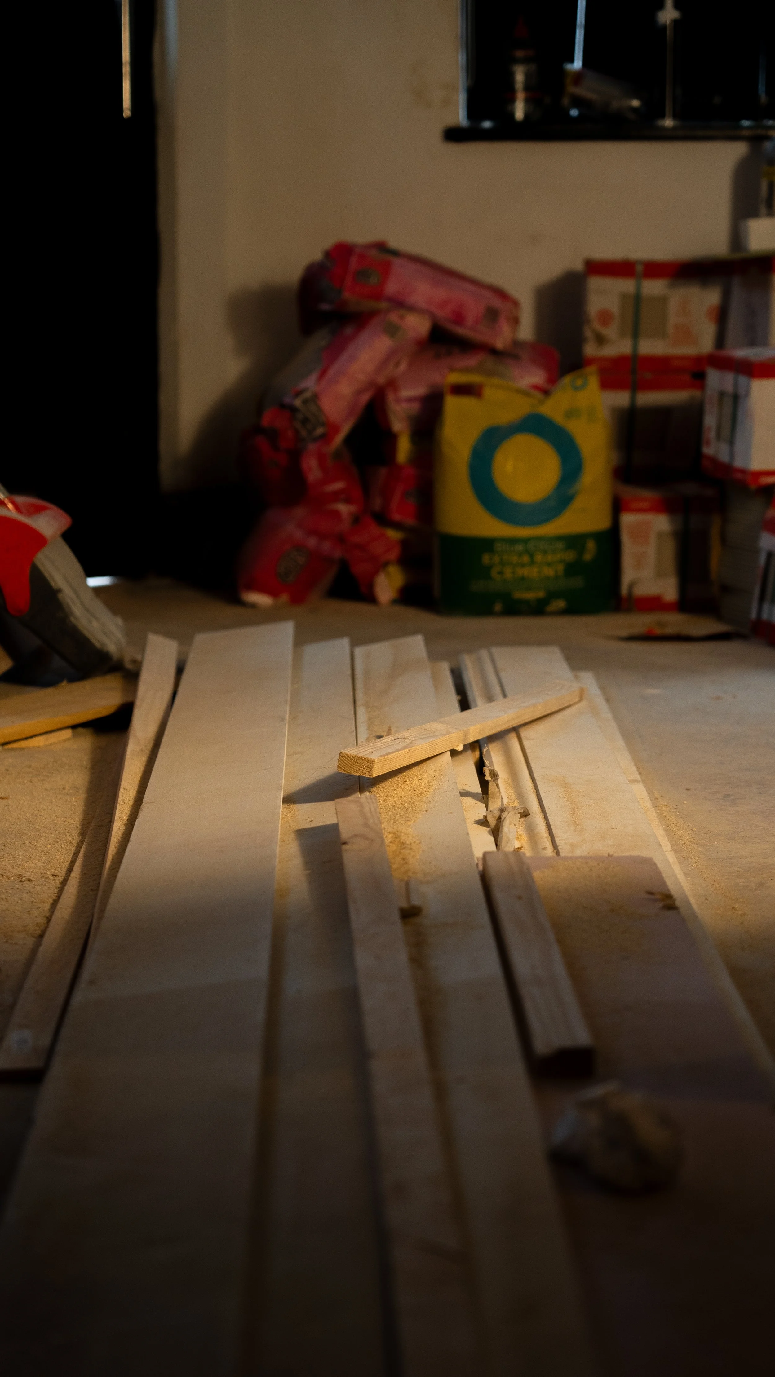 Wooden planks on a workbench with sawdust, with bags of cement and stored items in the background.