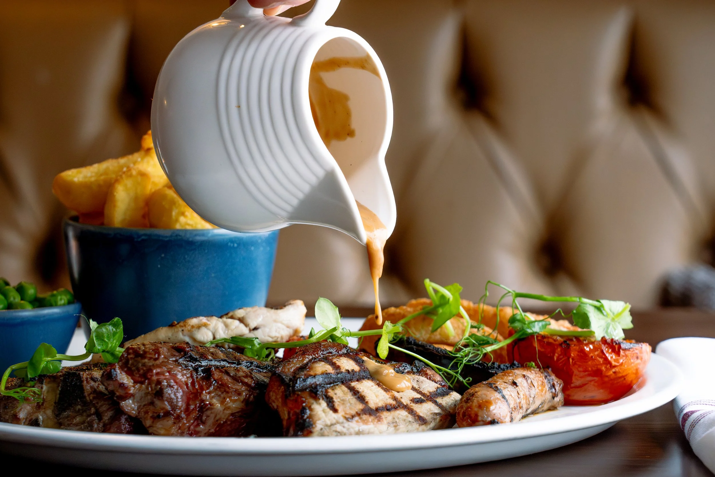 A plate of grilled meats, including steak, chicken, and sausage, with grilled vegetables and greens. A white gravy boat is pouring sauce over the food. In the background, there is a bowl of potato chips and a small bowl of peas.