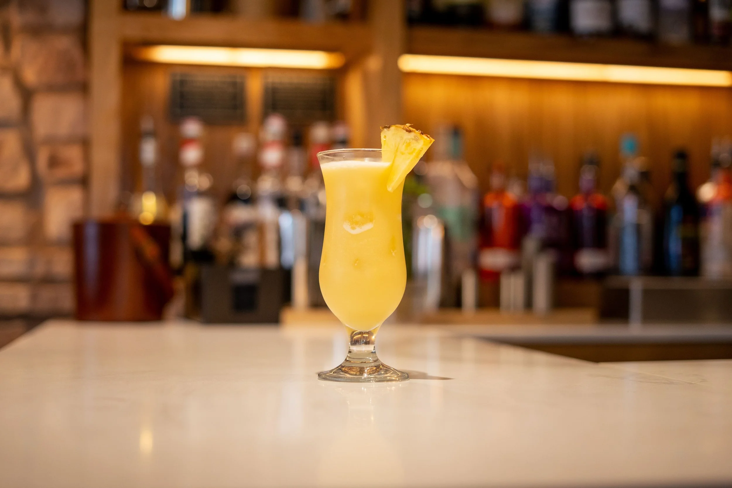 A tropical cocktail with pineapple garnish on a bar counter, with a blurred background of liquor bottles and wooden shelves.
