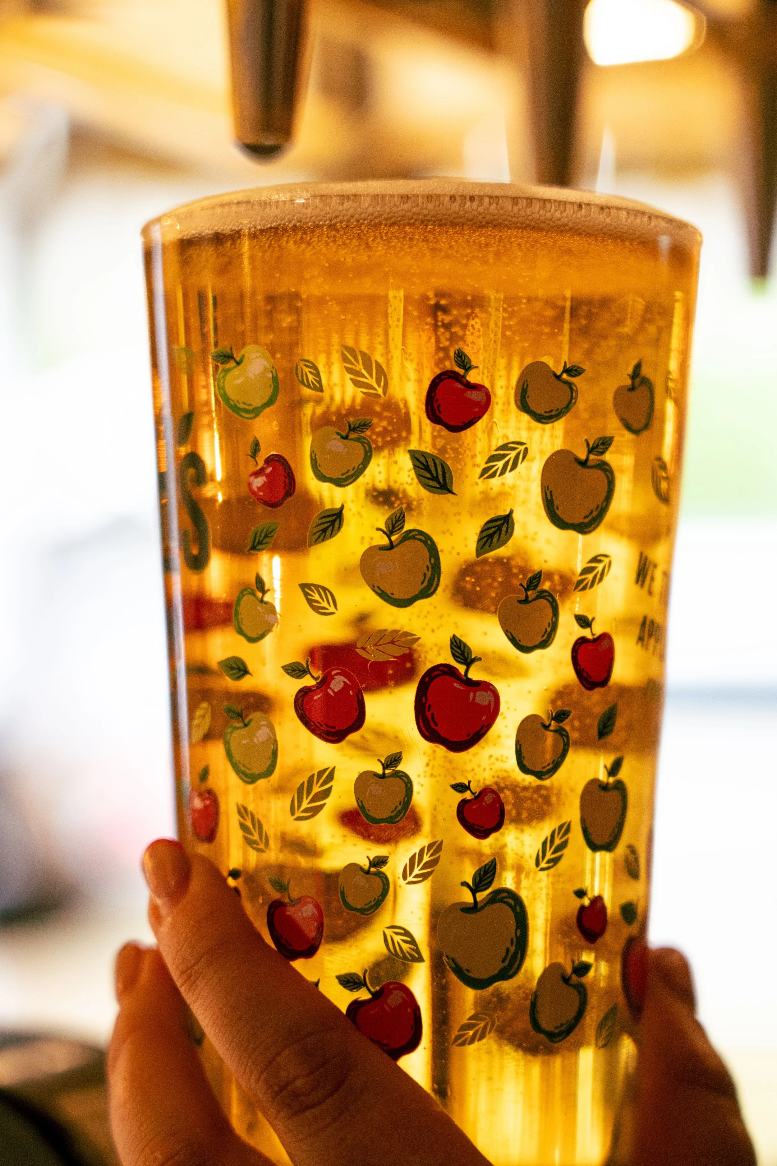 A hand holding a transparent yellow tumbler with apple and leaf stickers, filled with a bubbly amber-colored beverage.