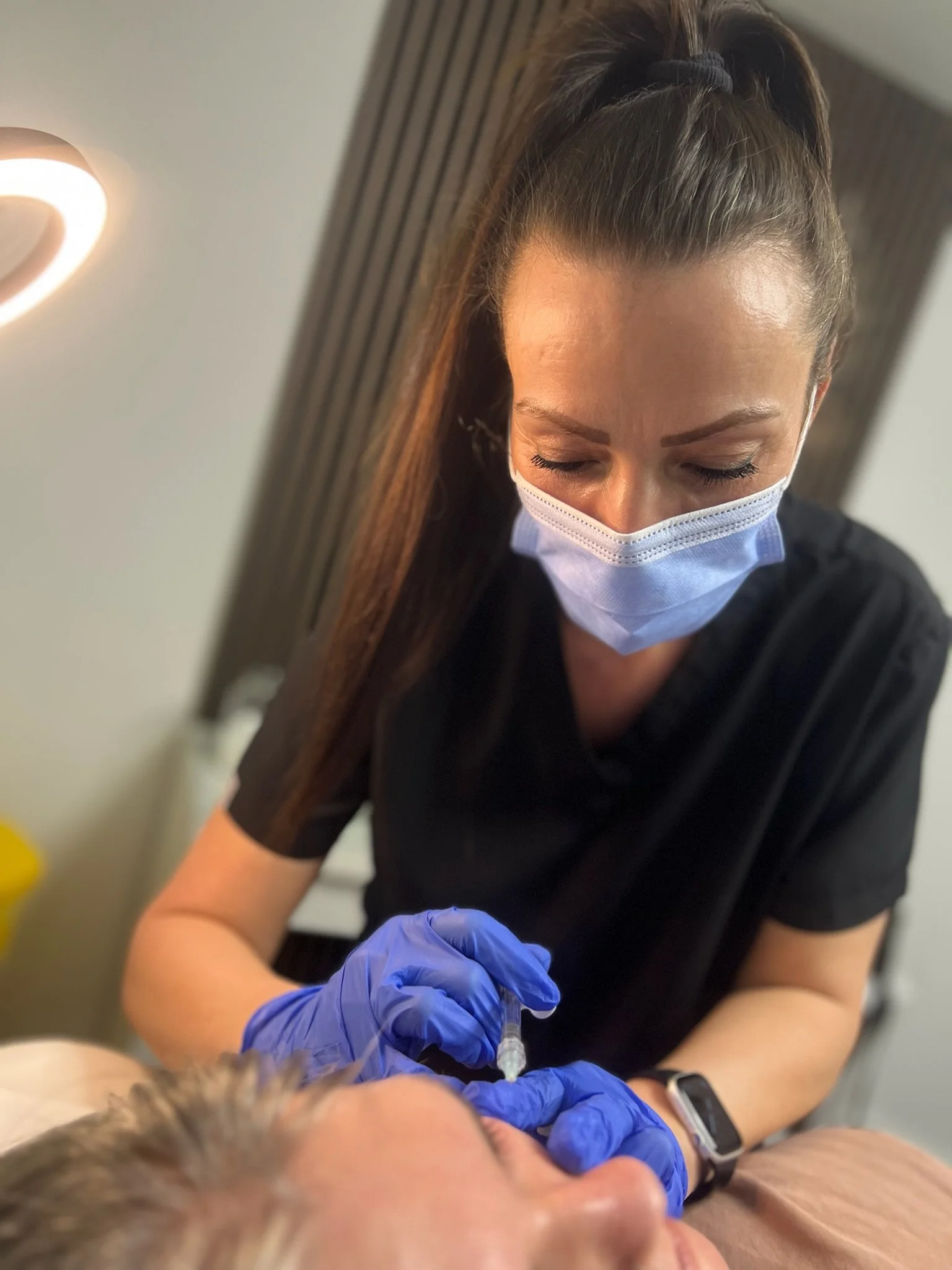 Elaine of Elaine Adams Aesthetics wearing a face mask and gloves is administering dermal filler to a client. The woman has long brown hair tied back and is wearing a black shirt and a smartwatch.