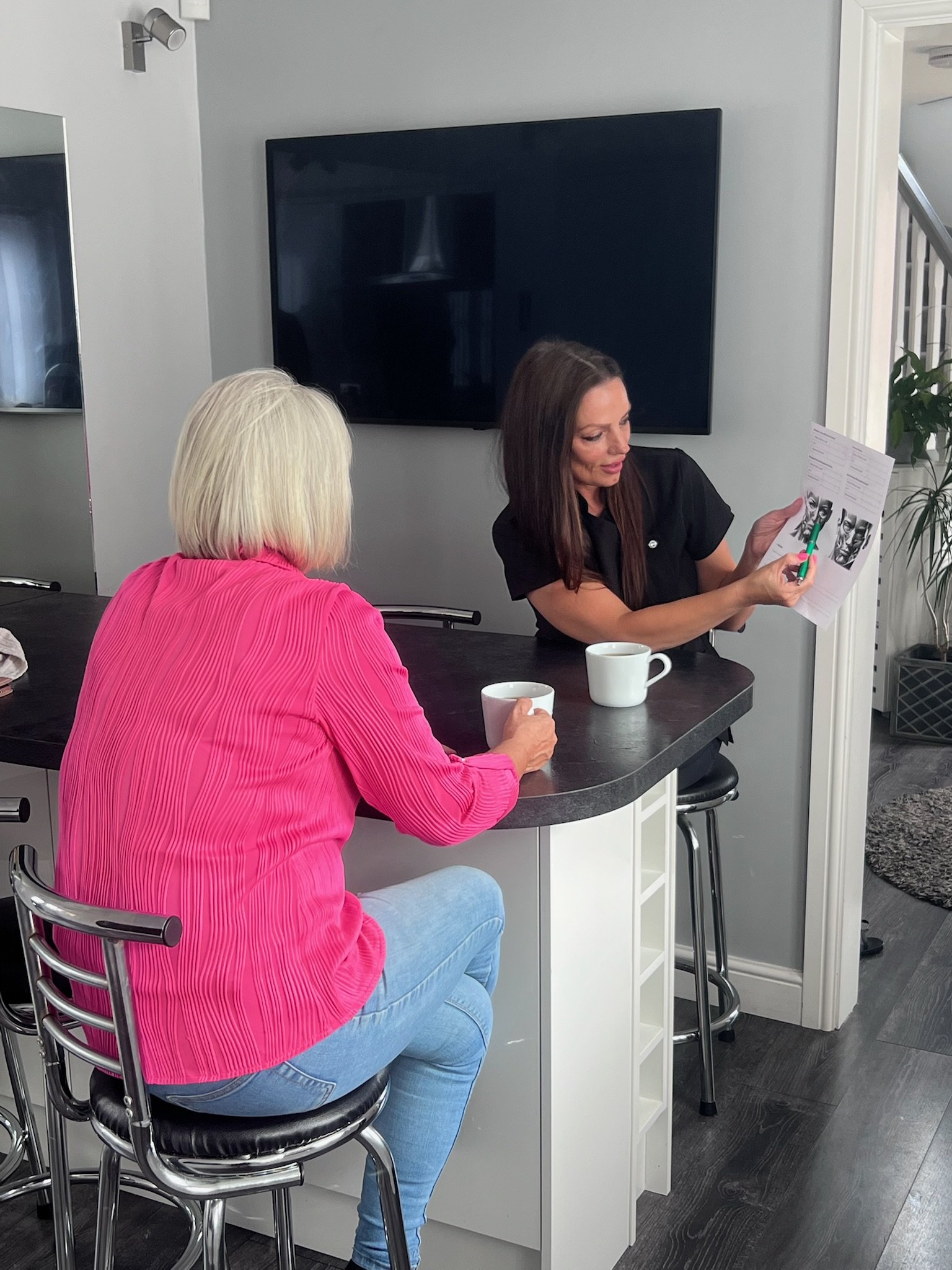Women sitting at a kitchen counter undertaking a consultation - one with platinum blonde hair wearing a bright pink jacket, the other with brown hair wearing a black top, holding a sheet of paper with photos, with coffee mugs on the counter.