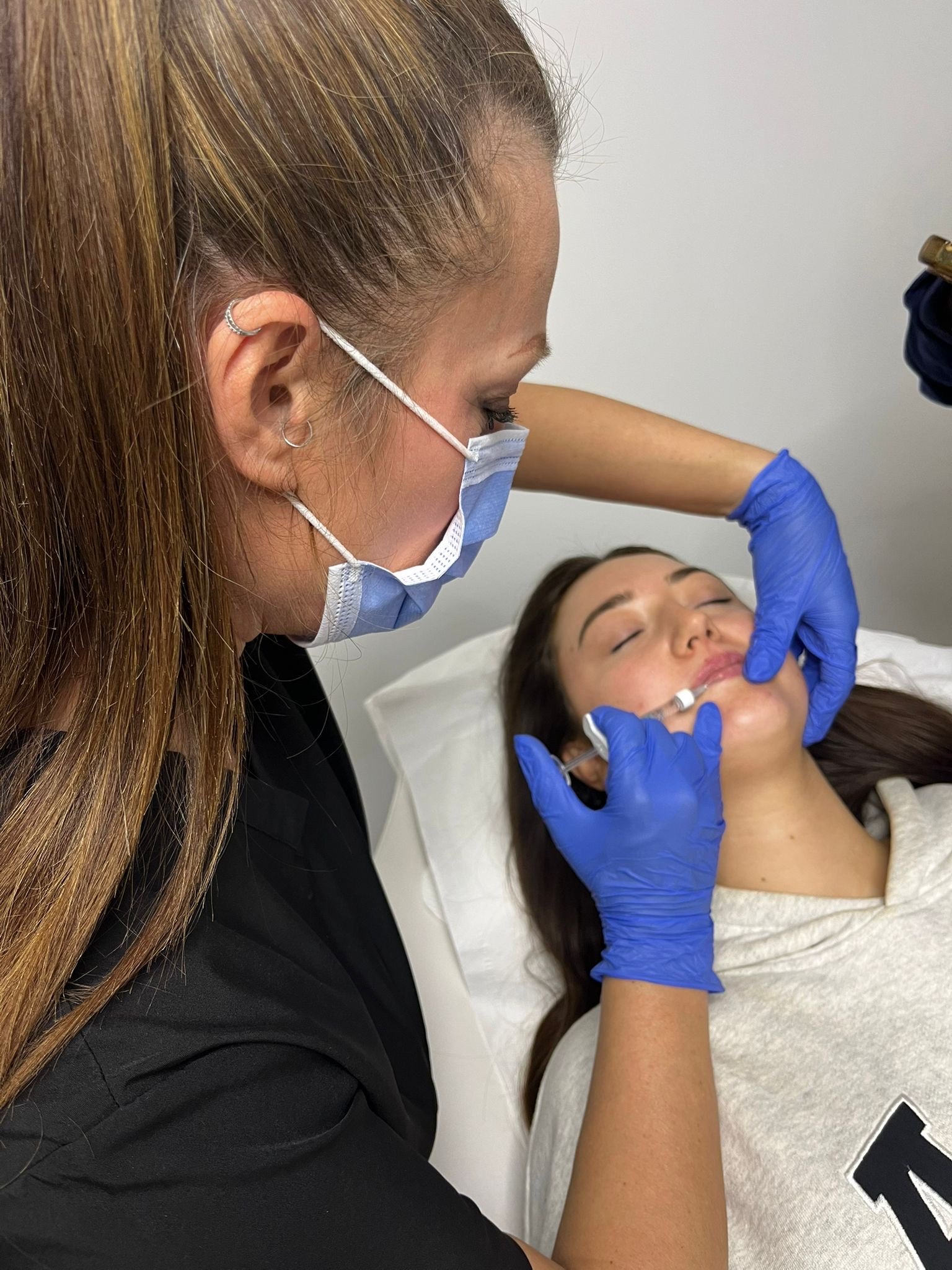 Medical professional (Elaine of Elaine Adams Aesthetics) administering an injection to a woman lying on a treatment bed in a clinical setting.