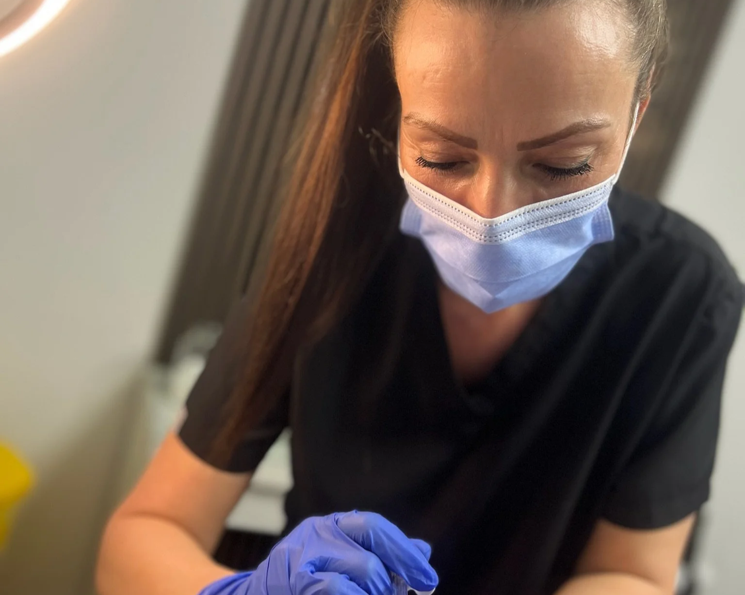 Elaine of Elaine Adams Aesthetics wearing a face mask and gloves performing a cosmetic procedure, indoors.