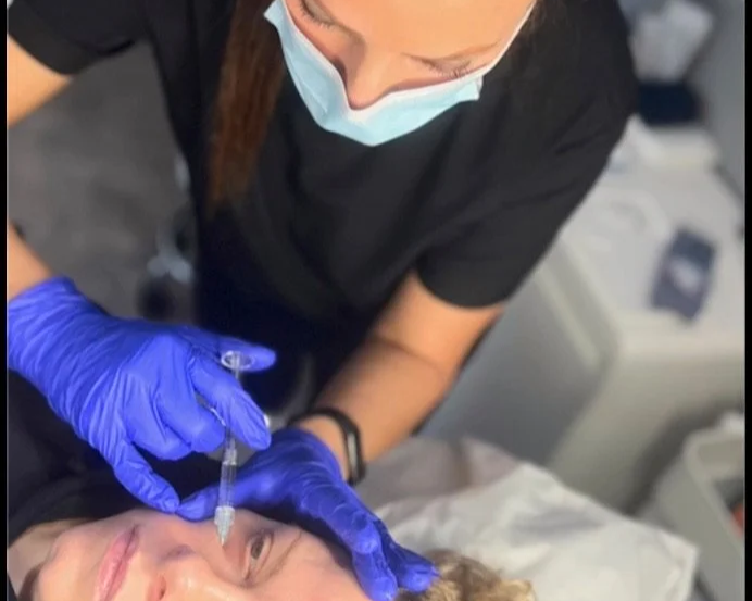 Elaine of Elaine Adams Aesthetics administering a facial injection to a patient lying down, wearing gloves and a face mask.