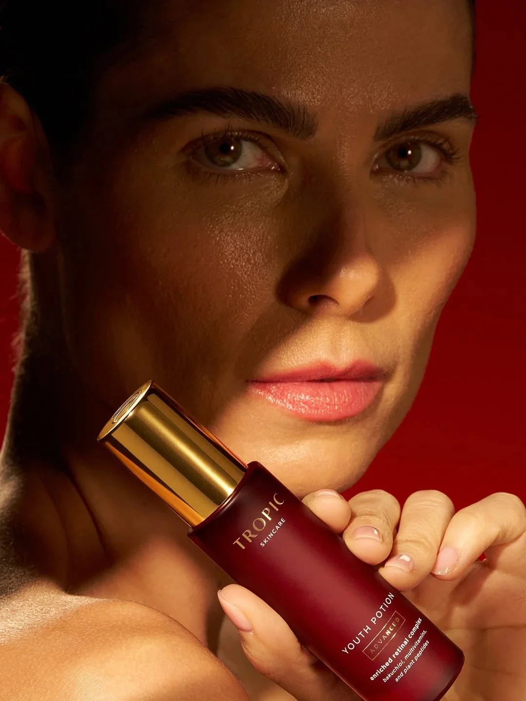 A close-up of a woman holding a bottle of Tropic Skincare Youth Potion. She has clear skin, dark eyebrows, and is smiling slightly. The background is red.