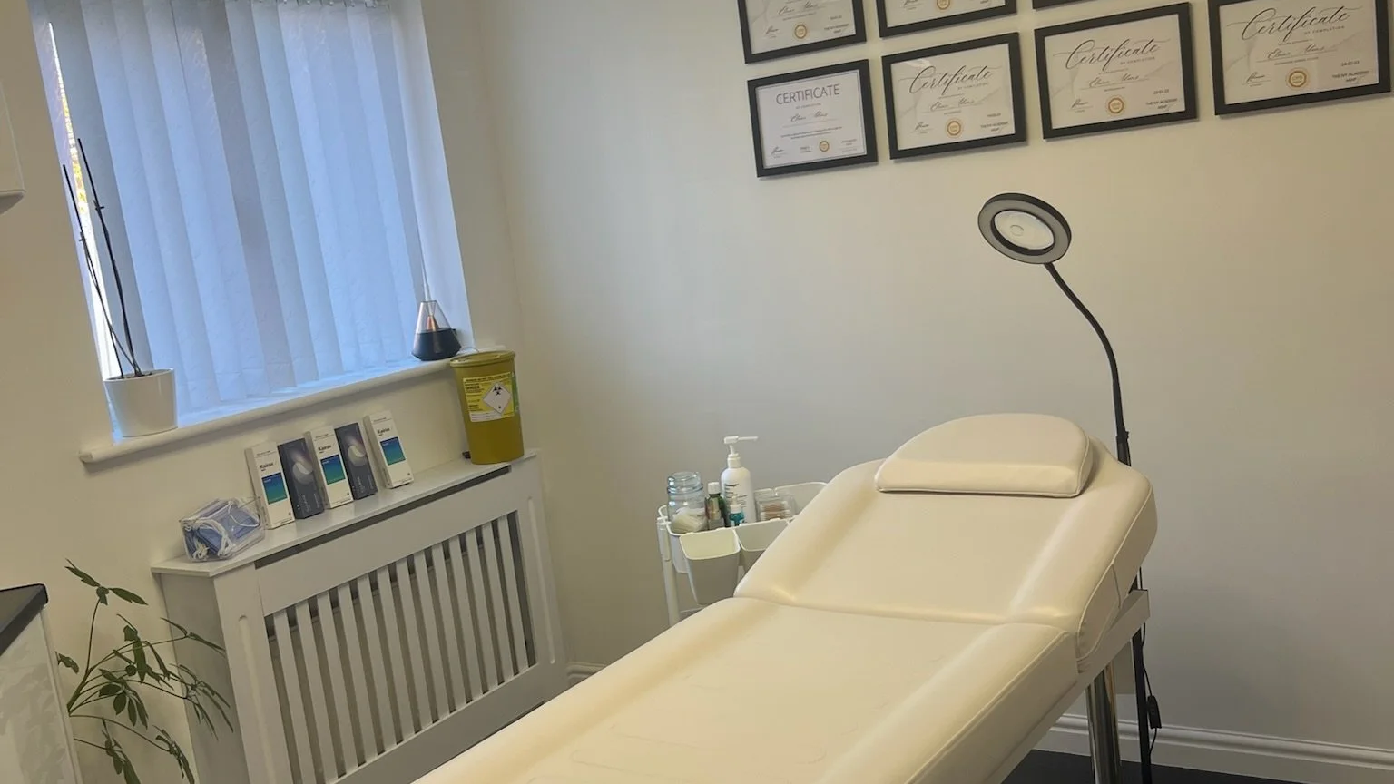 The Elaine Adams Aesthetics treatement room in Scissett, Huddersfield with a cream-colored examination table, framed certificates on the wall, sunlit window with blinds, and various medical supplies including hand sanitizer biohazard container.