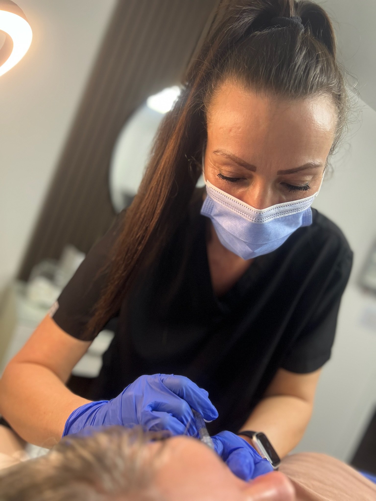 Elaine of Elaine Adams Aesthetics is administering an injectable cosmetic treatment to a client. The woman has long brown hair tied back and is focused on her task.