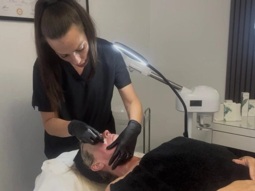 Elaine of Elaine Adams Aesthetics performing a facial treatment or skincare procedure on a client lying on a bed, using a handheld device while wearing black gloves.