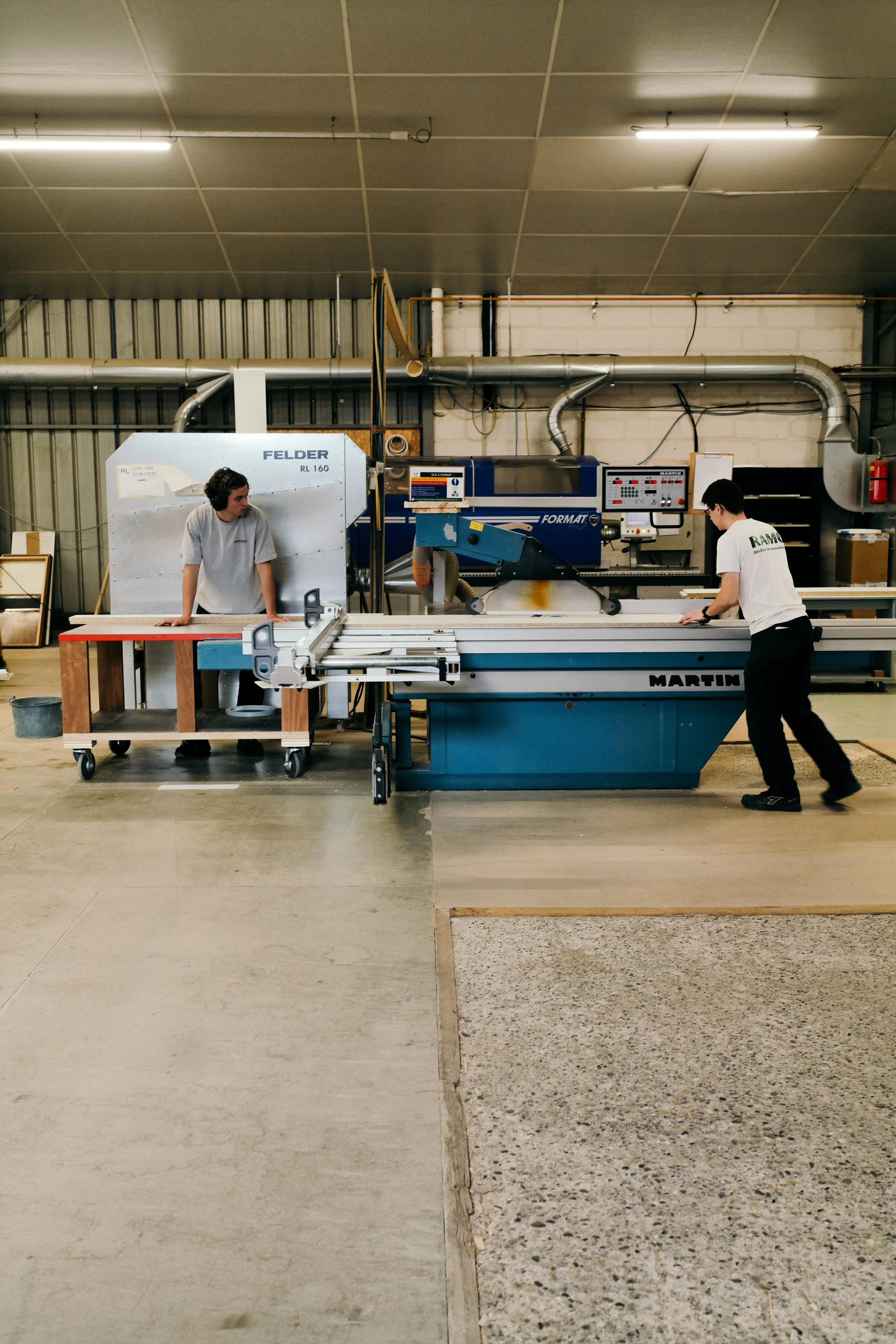 Deux hommes travaillant dans une usine sur une machine industrielle, l'un porte un t-shirt blanc et l'autre un t-shirt gris, le premier porte des lunettes.