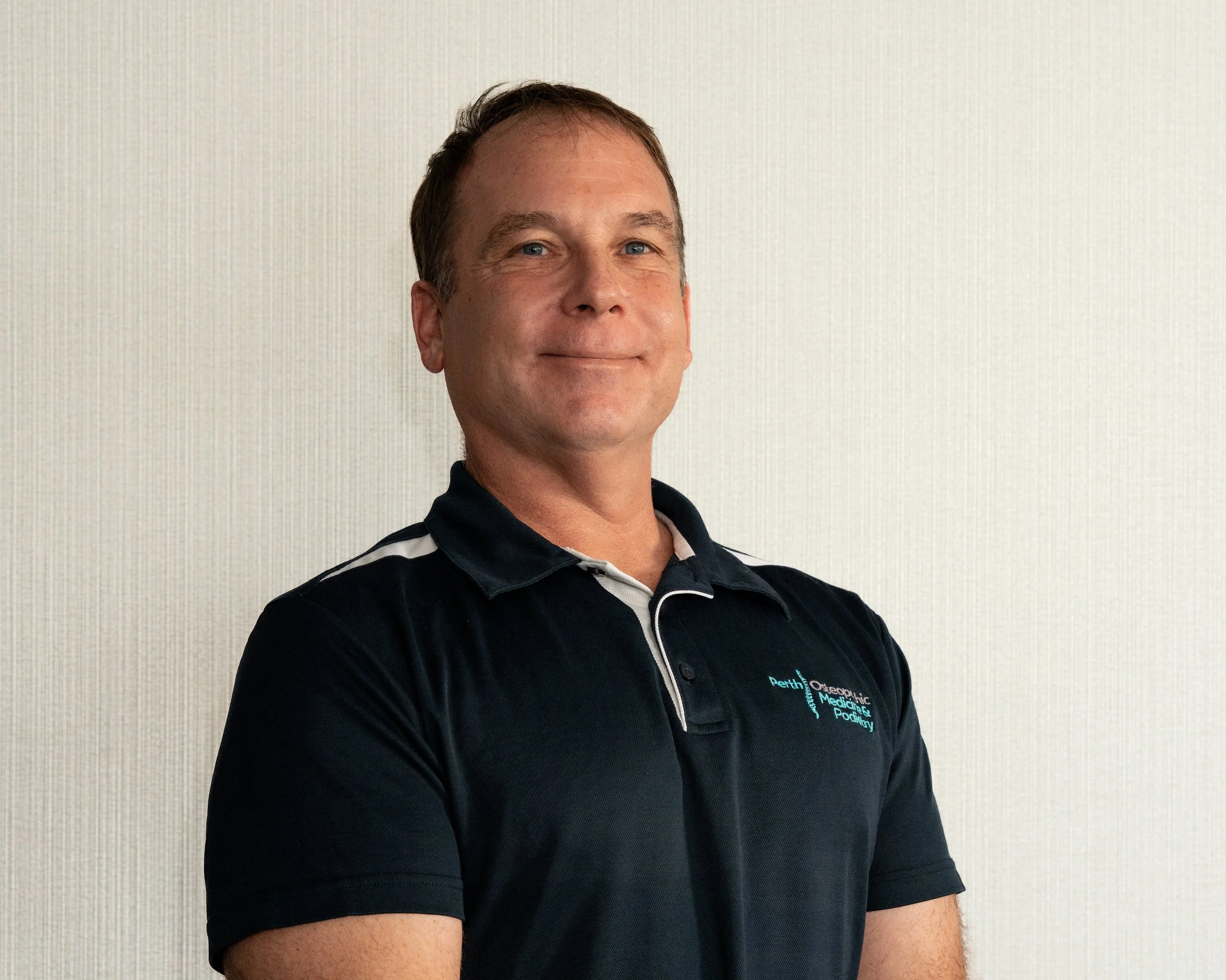 Osteopath Dr Denis Winter - Owner of Perth Osteopathic Medicine & Podiatry posing in a branded Perth Osteo collared shirt.