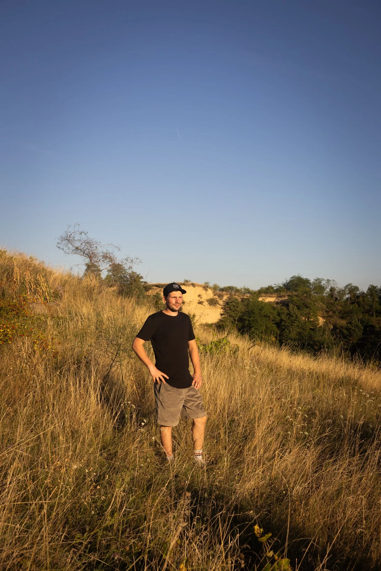 Arnold Holzer steht in einer offenen Wagramer Graslandschaft bei Sonnenuntergang, trägt ein schwarzes T-Shirt, Bermudashorts und eine schwarze Kappe, im Hintergrund sind Bäume und Hügel zu sehen.