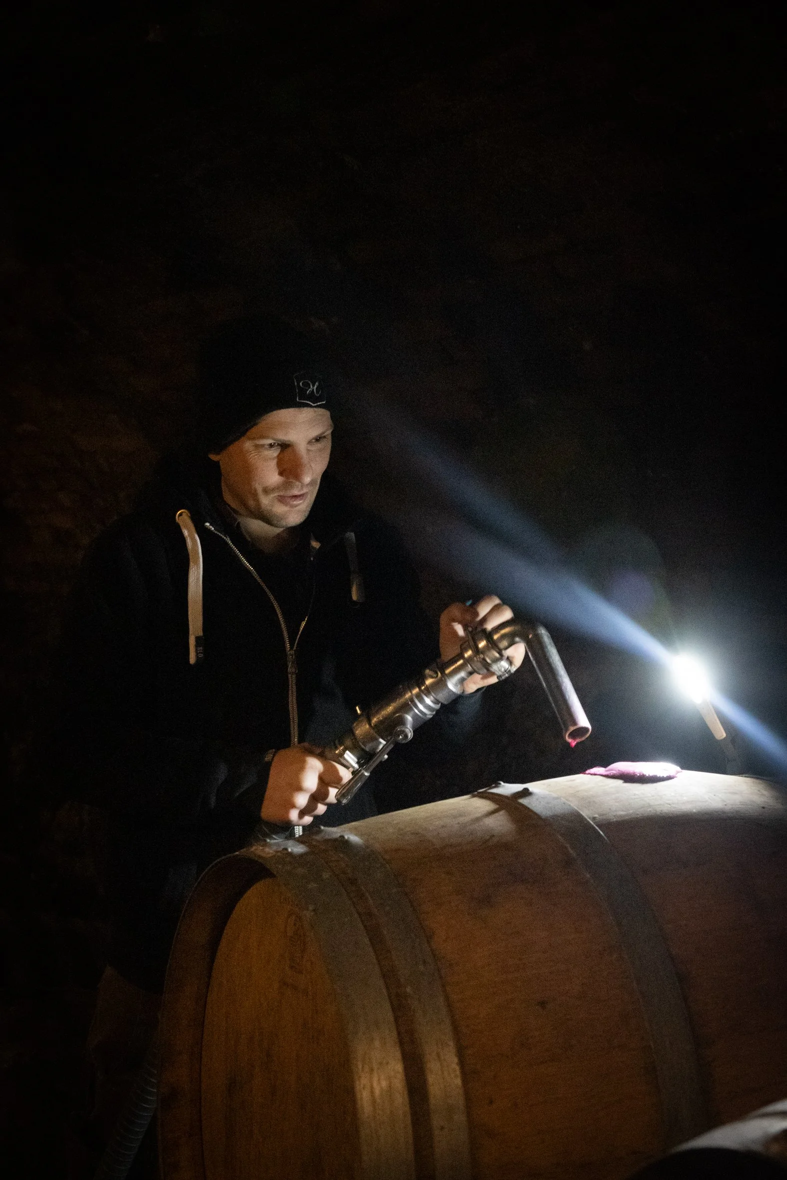 Ein Mann trägt eine schwarze Mütze und eine schwarze Jacke. Er hält eine Taschenlampe und leuchtet auf eine Holzscheibe, während es dunkel ist.