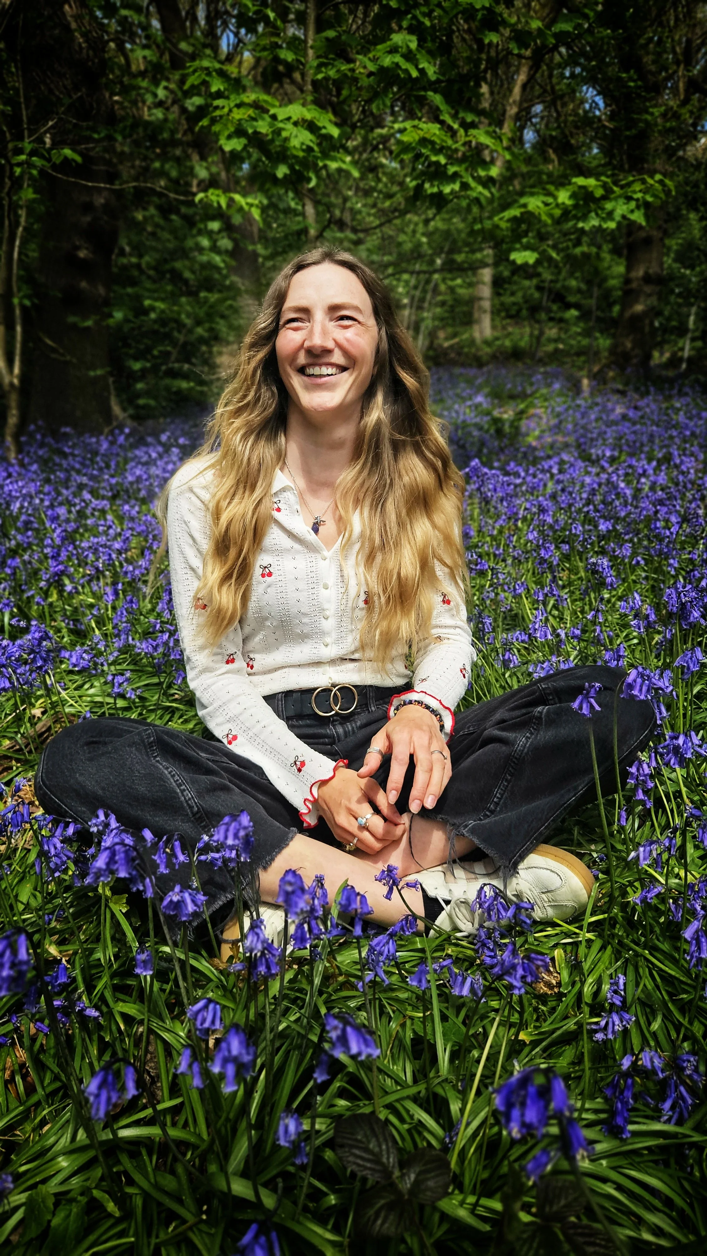 A woman with long, wavy blonde hair sitting cross-legged in a field of purple flowers, smiling and looking to her right. She is wearing a white shirt with small red and black accents, black jeans with frayed hems, and white sneakers. The background features green trees and foliage.