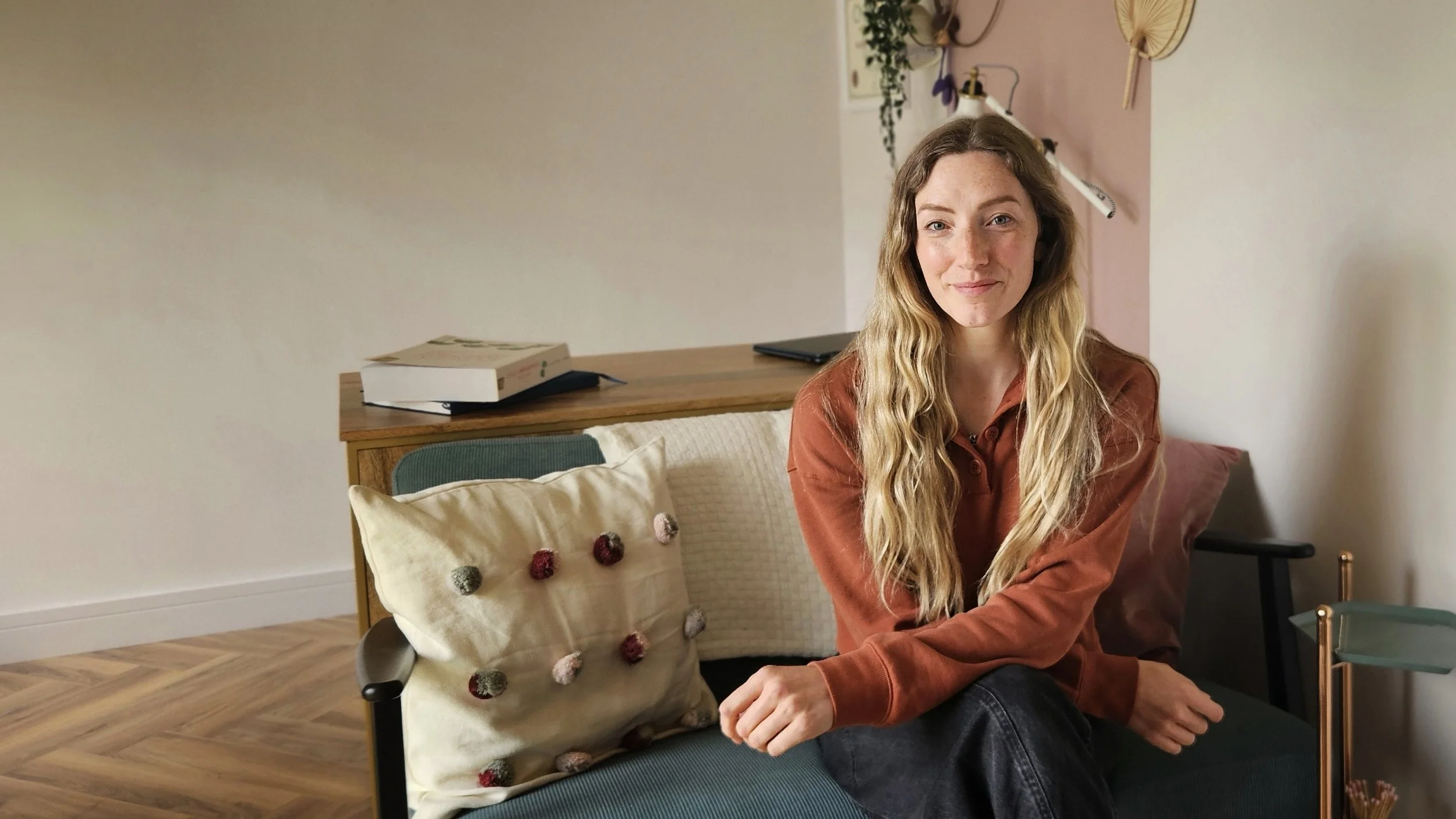 A young woman with long wavy blonde hair and freckles sitting on a chair in a cozy room, wearing an orange-brownish long-sleeve shirt. Behind her, a wooden side table with books, and a wall decorated with plants, fans, and wall art. A decorative pillow with pom-poms is on the chair.