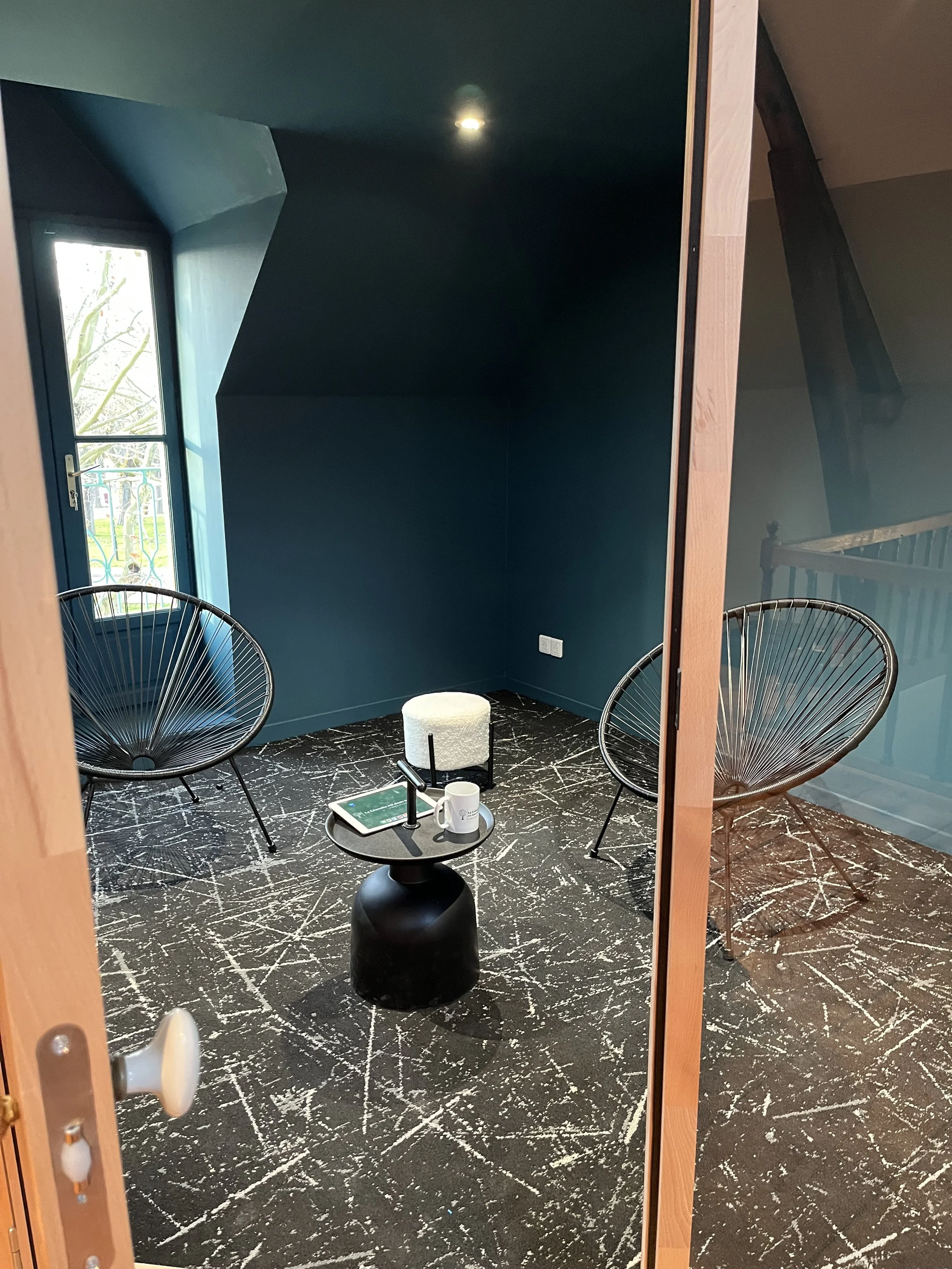 Une pièce de style moderne avec deux chaises en forme de rayon de soleil en acier noir, un petit tapis blanc, une table noire avec une tasse blanche et une tablette, murs bleu foncé et sol en marbre noir avec des veines blanches, une fenêtre laissant entrer de la lumière naturelle.